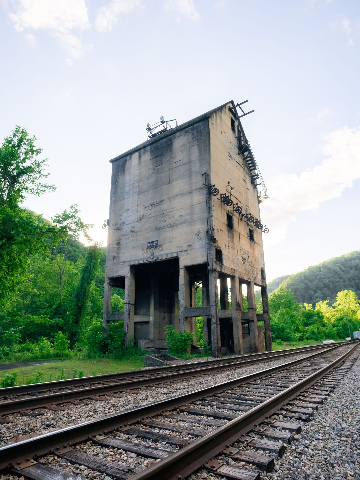 Abandoned concrete coal tower stands alongside railroad tracks in a lush green forest setting.