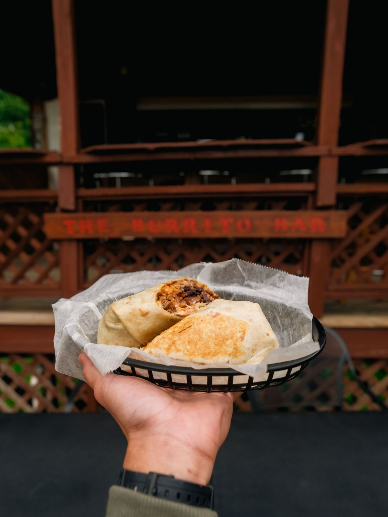 Hand holding a grilled sandwich served in a basket with parchment paper at an outdoor restaurant setting.