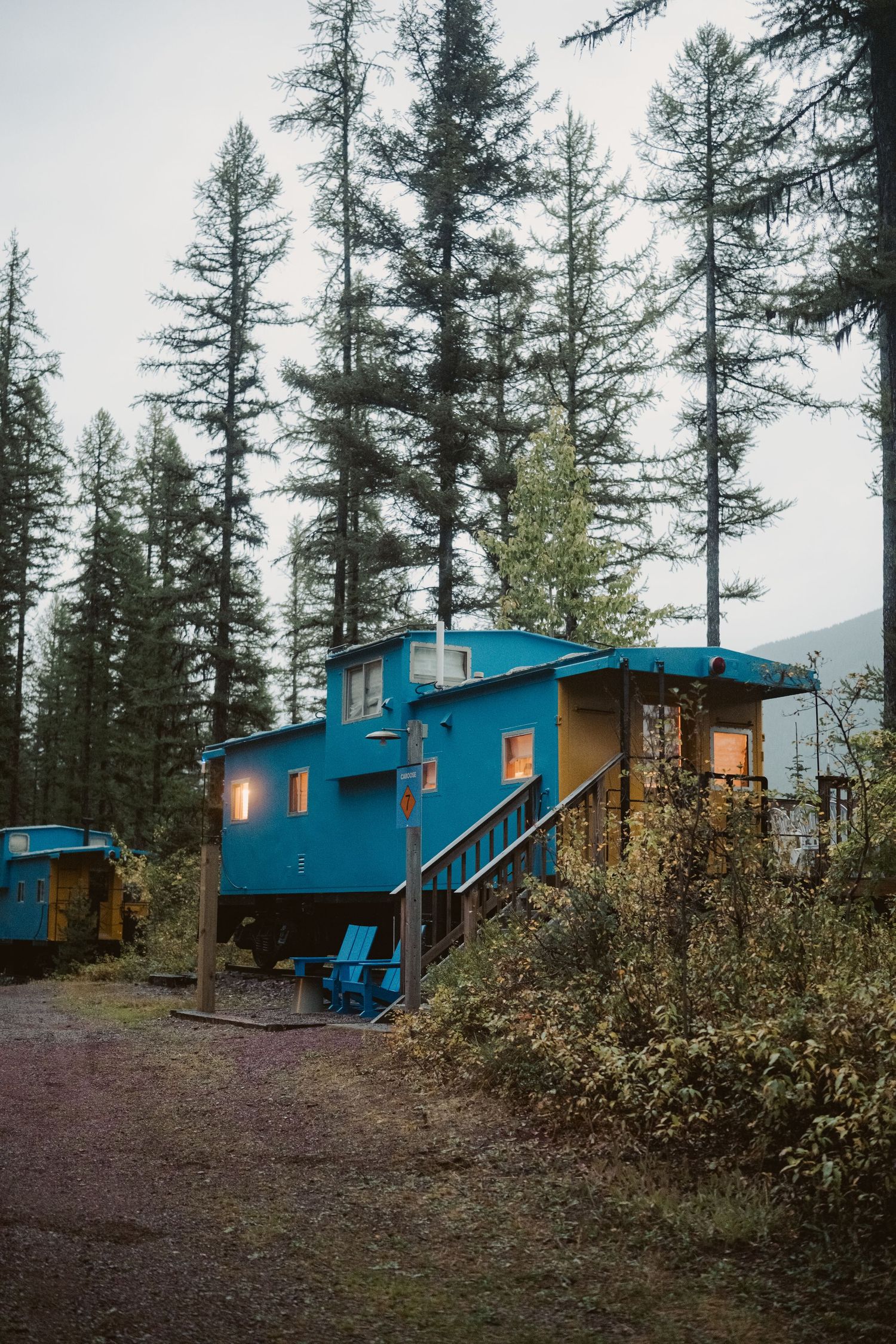 Rustic blue cabin lodges nestled among tall evergreen trees at dusk in a remote mountainous setting.