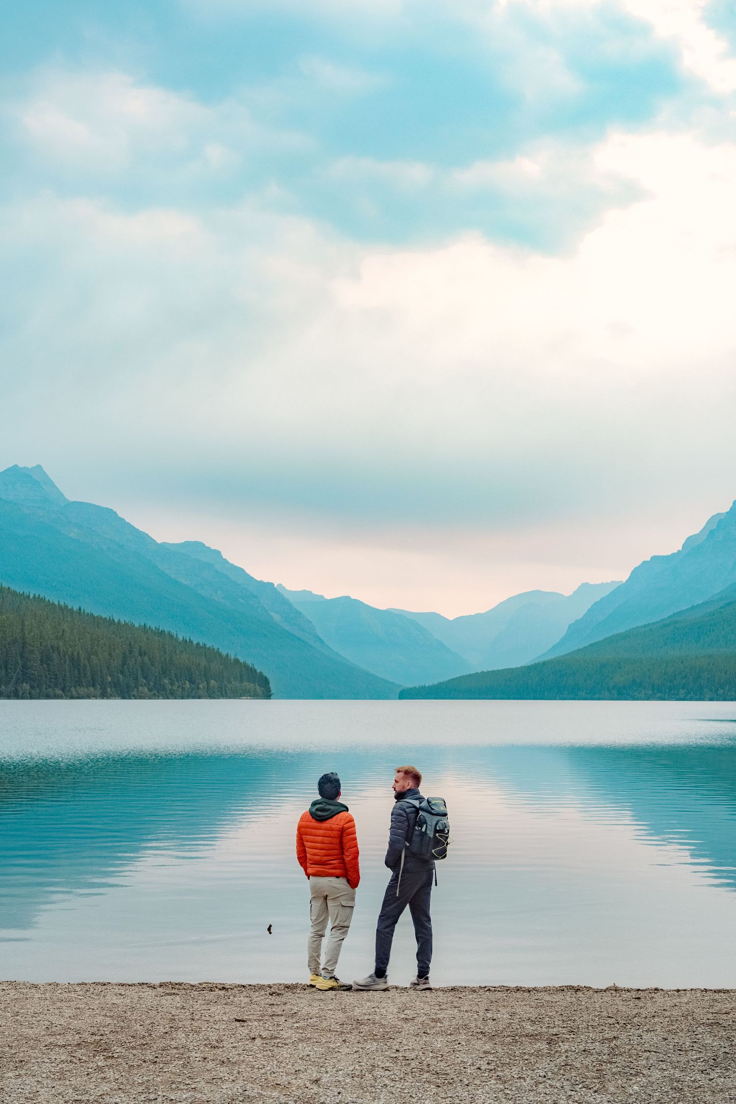 Two people admiring the tranquil waters and mountain views at a serene lakeside beach in Glacier National Park.