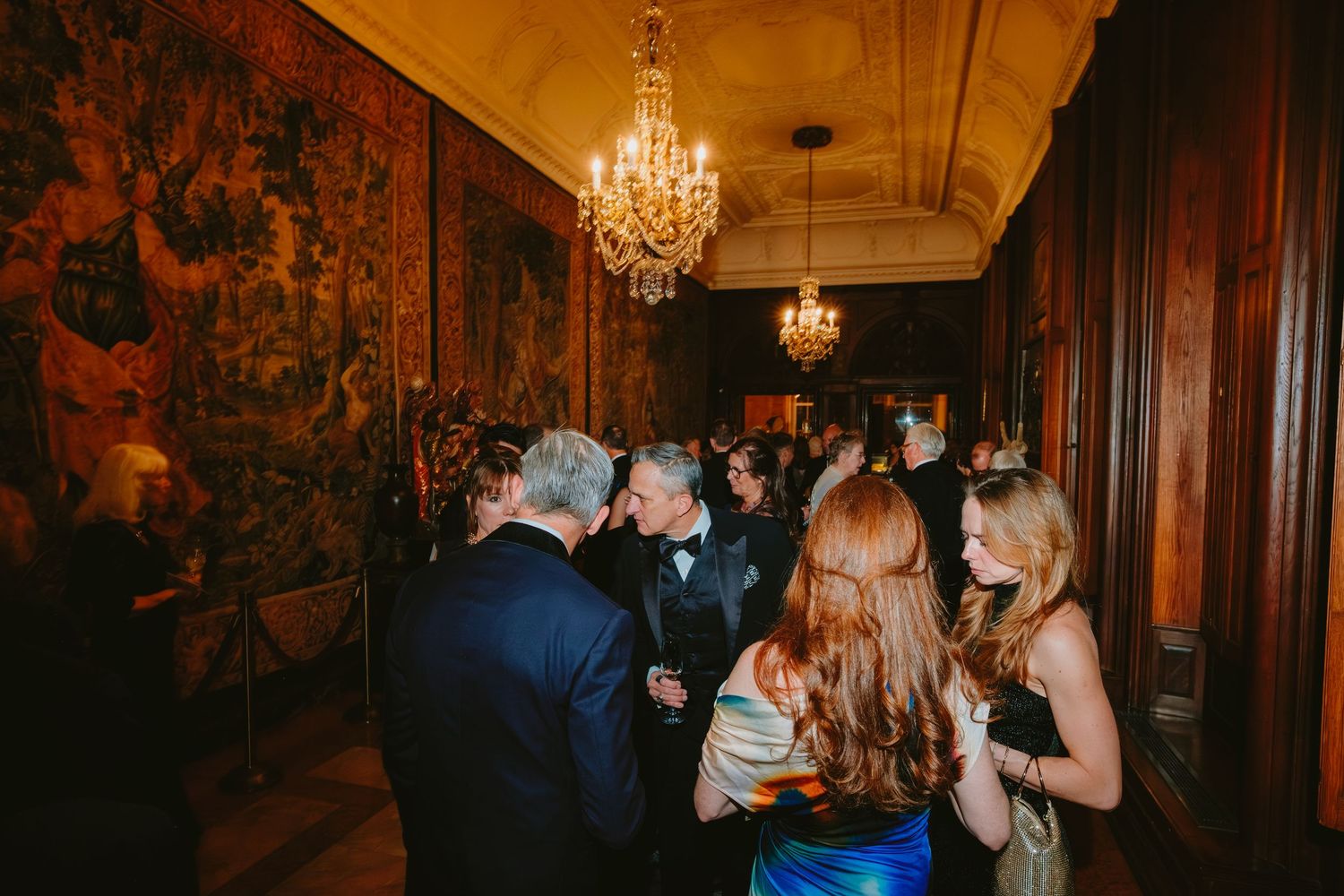 A couple dances together under an elegant crystal chandelier in a vintage-style ballroom with dark wood paneling.