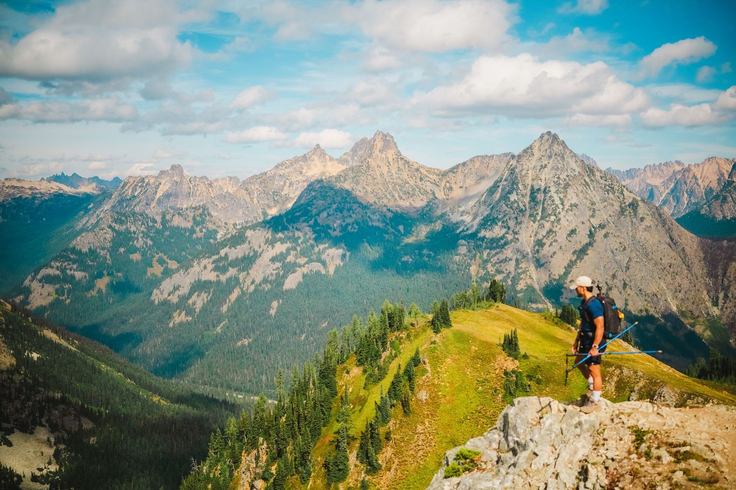 Hiker stands on mountain summit overlooking dramatic jagged peaks under blue cloudy sky.