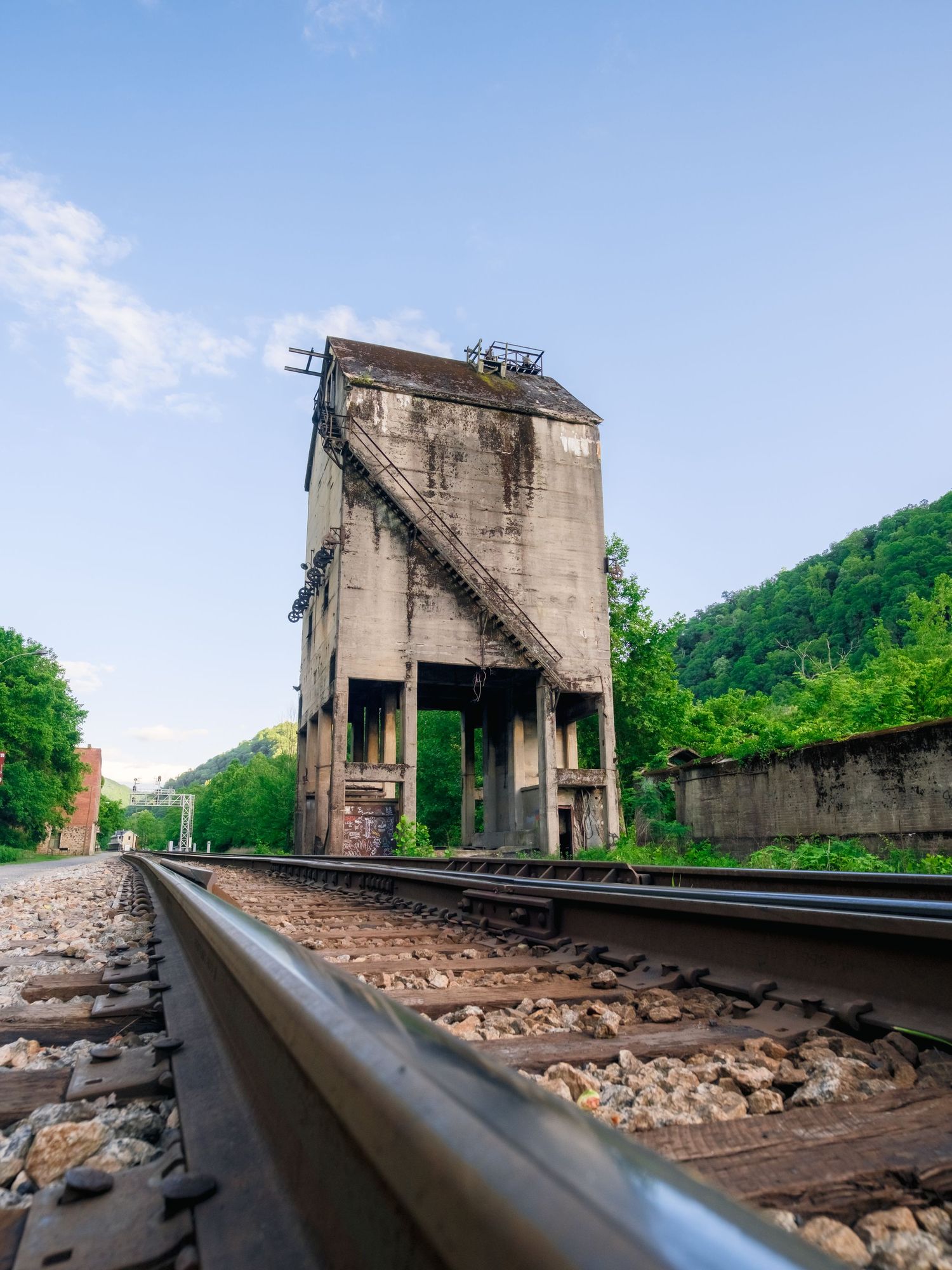 Abandoned concrete railroad coaling tower looms over train tracks in a green mountainous landscape on a sunny day.