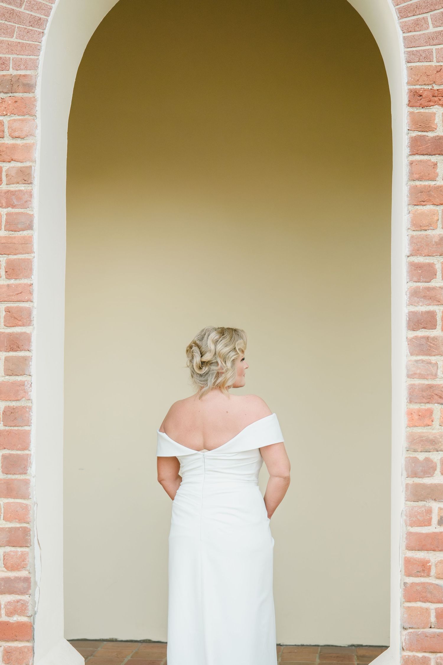 Back view of a bride in an off-shoulder white wedding gown standing in a brick archway.