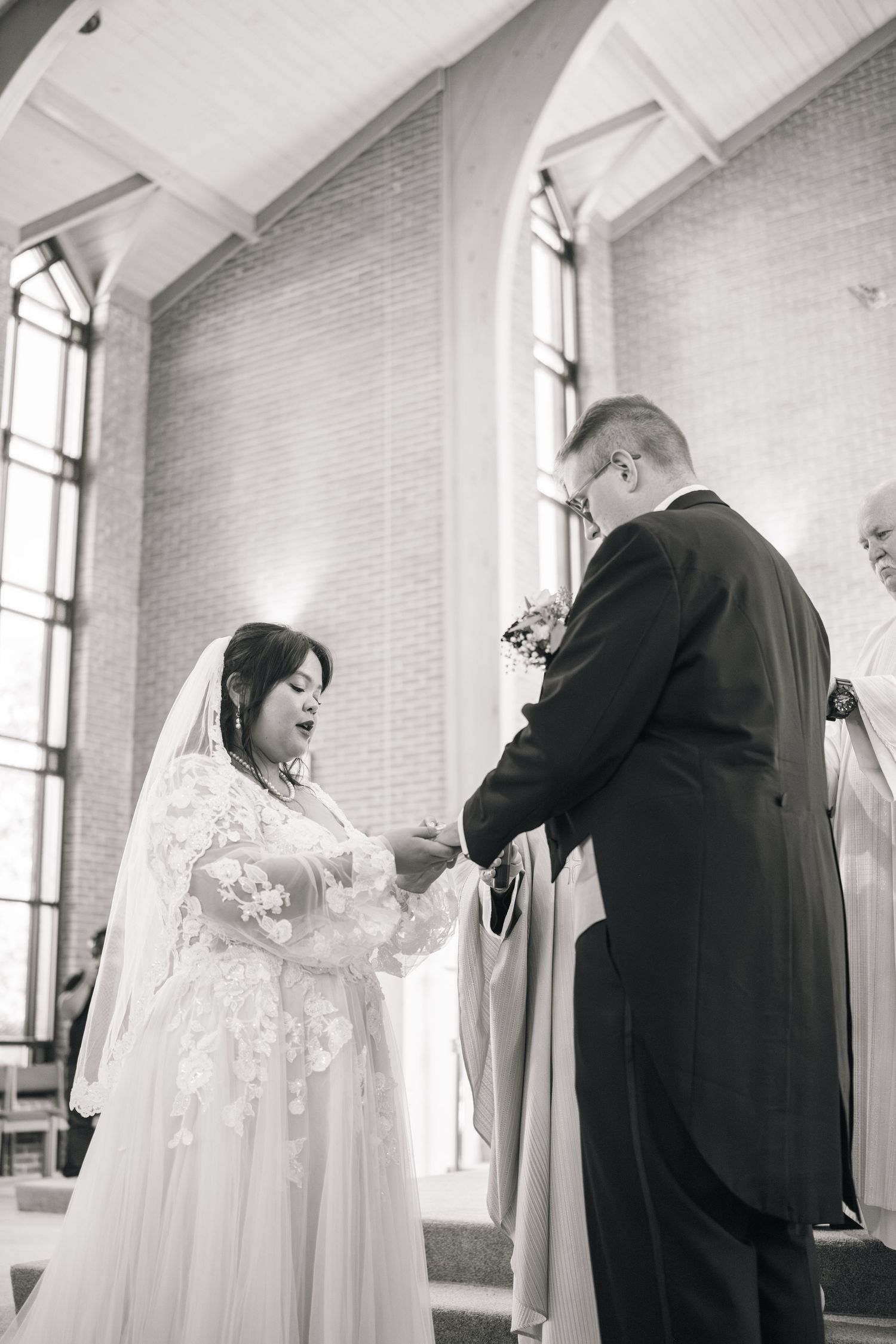A couple exchanges rings during their wedding ceremony in a church with tall windows in black and white photography.