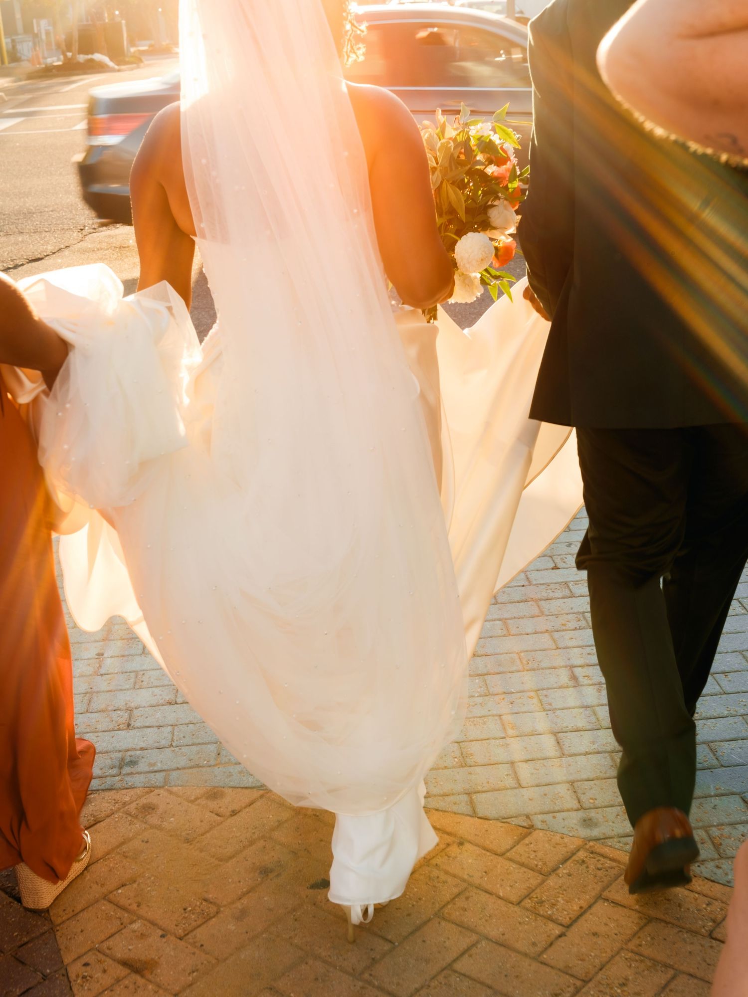 Flowing white wedding dress train being carried across brick pavement in golden sunlight.