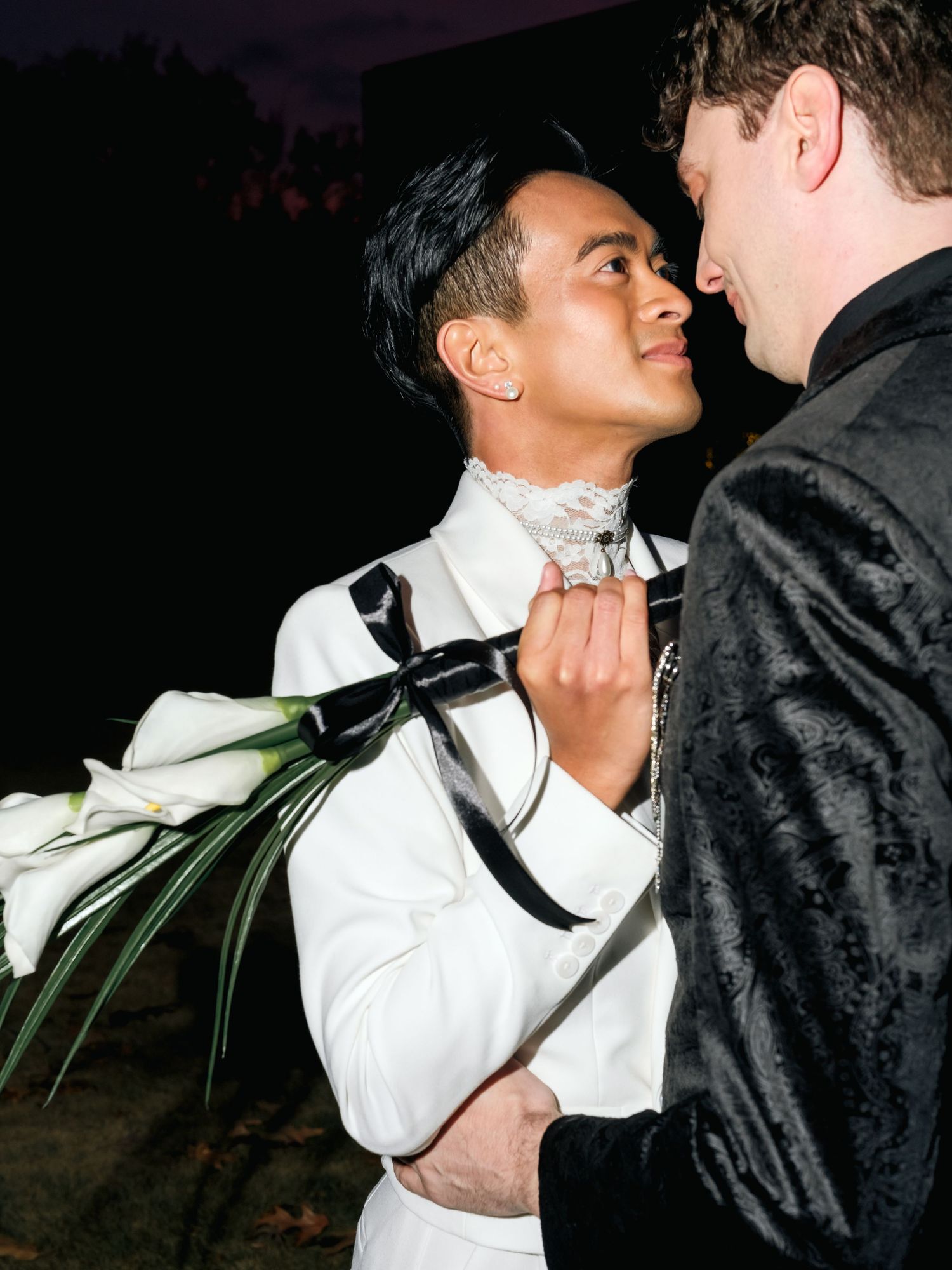 Two people share an intimate moment at a wedding celebration, with one wearing a white suit and holding a floral bouquet.