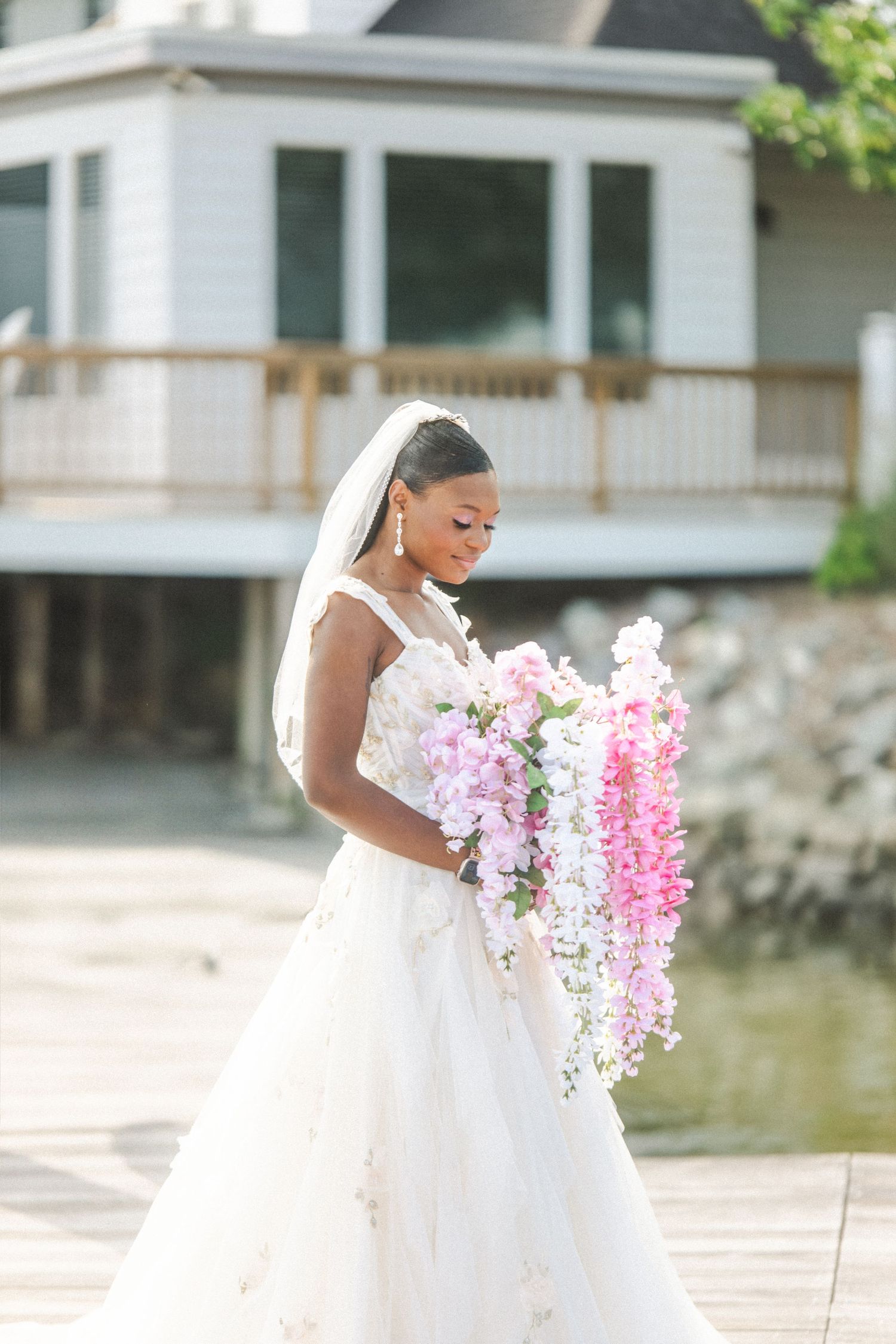 A bride in a white wedding dress holds a beautiful cascade of pink and white delphinium flowers on a lakeside dock.