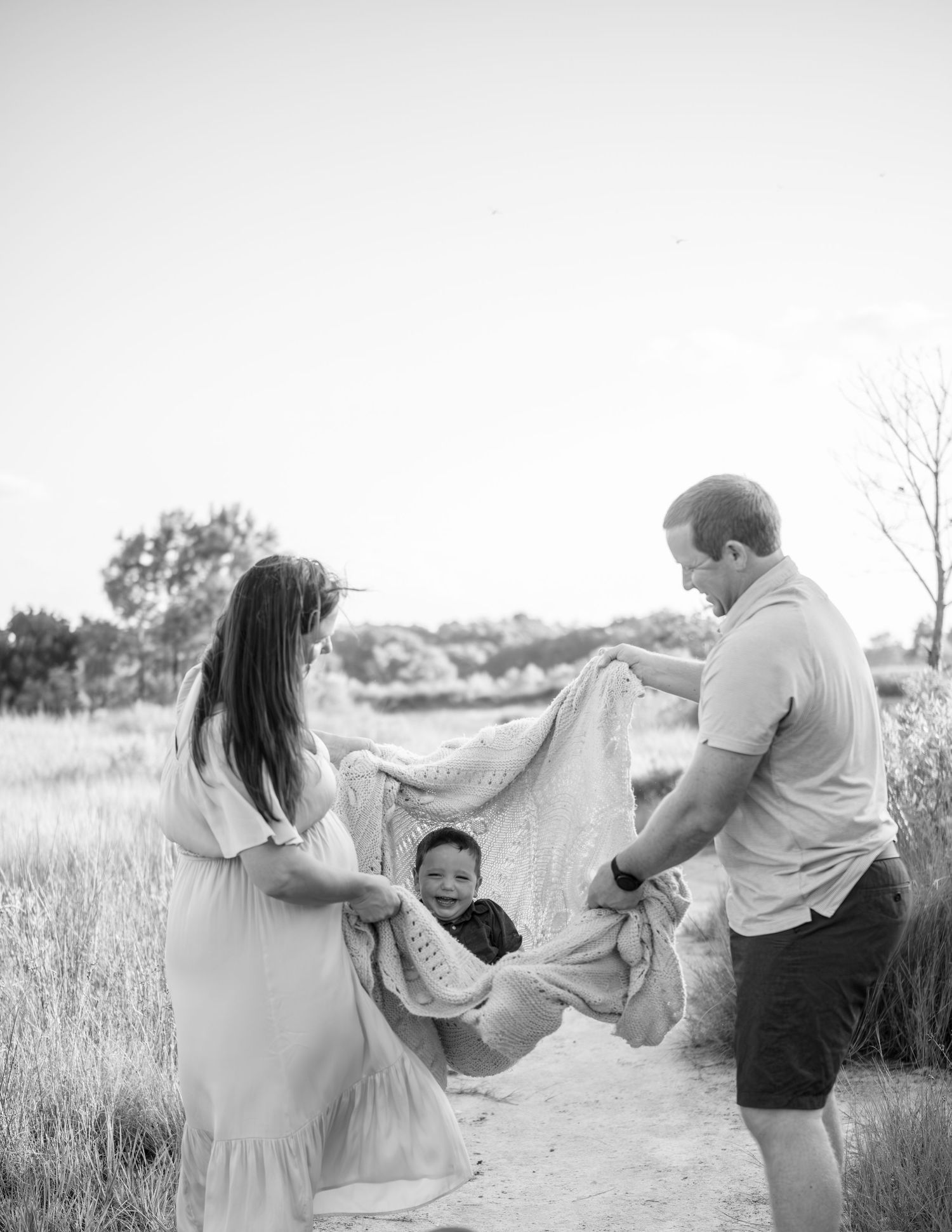 Family plays together in a grassy field, swinging a blanket with joy in black and white photography.