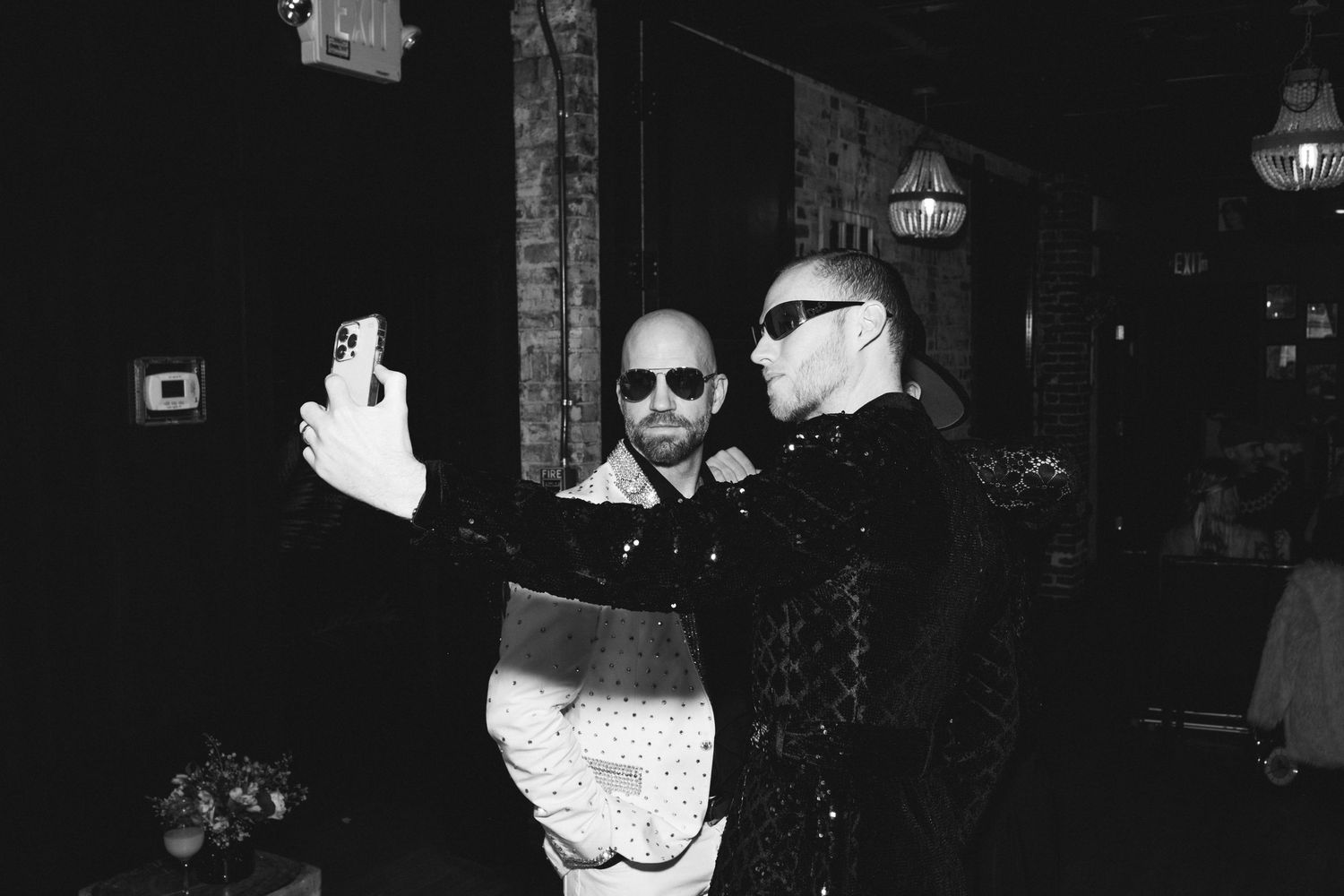 A black and white photo of two people in dark clothing taking a selfie together at a dimly lit venue.