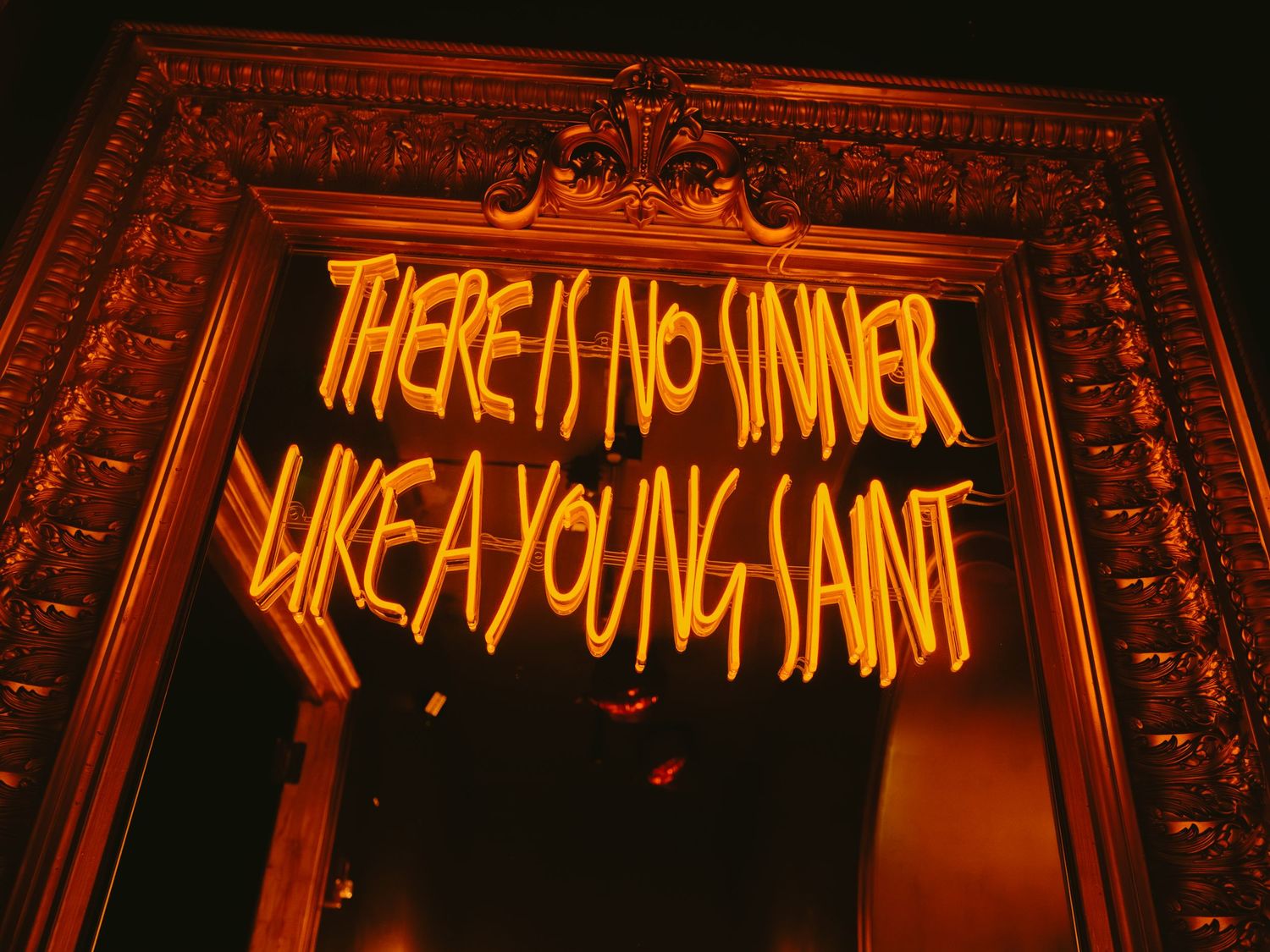 Neon orange text reading 'There is no sinner like a young saint' glows against a dark ornate frame.