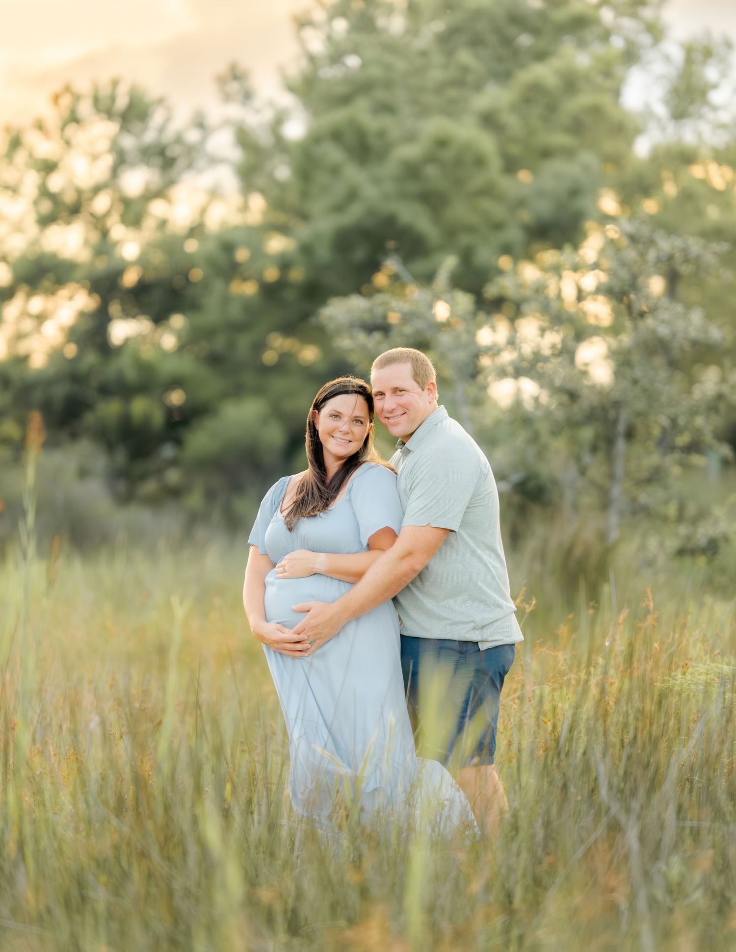 Couple embraces in golden field at sunset as they capture maternity photos in a dreamy outdoor setting.
