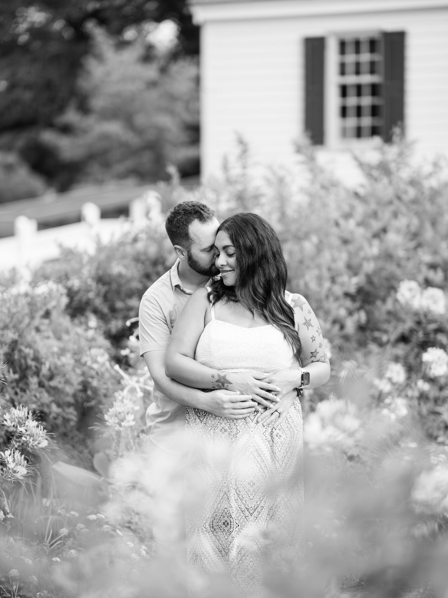 Romantic black and white photo of a couple embracing in a flowering garden with a white house in the background.