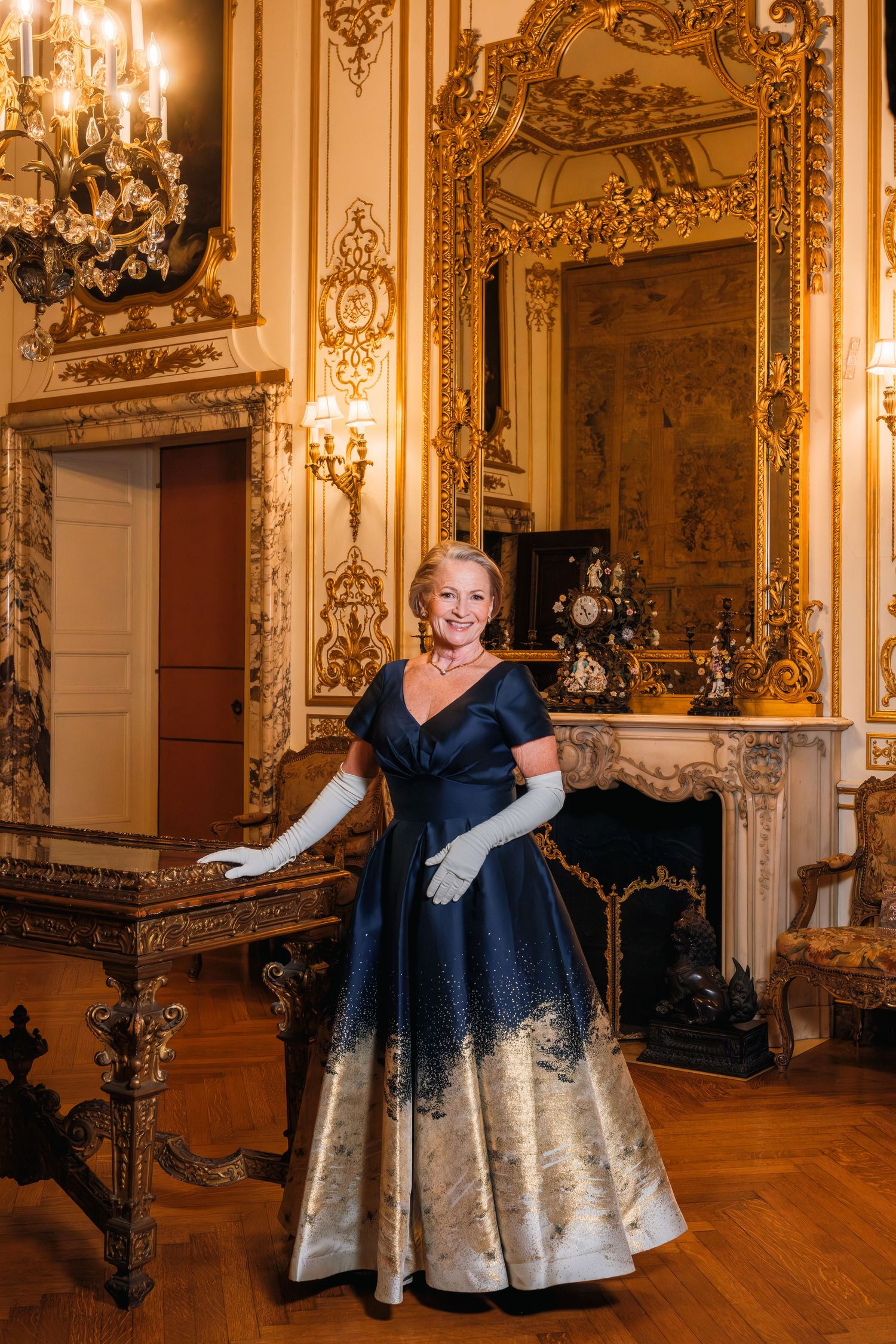 A person in an elegant blue and silver ballgown twirls gracefully across a grand ballroom with ornate gold decor.