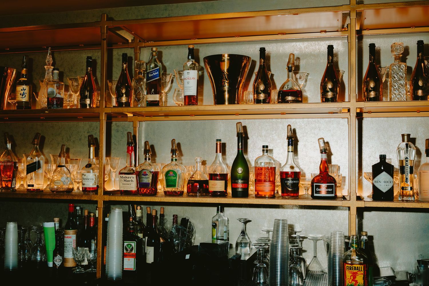 Wooden bar shelves stocked with various liquor bottles and glasses illuminated by warm lighting.