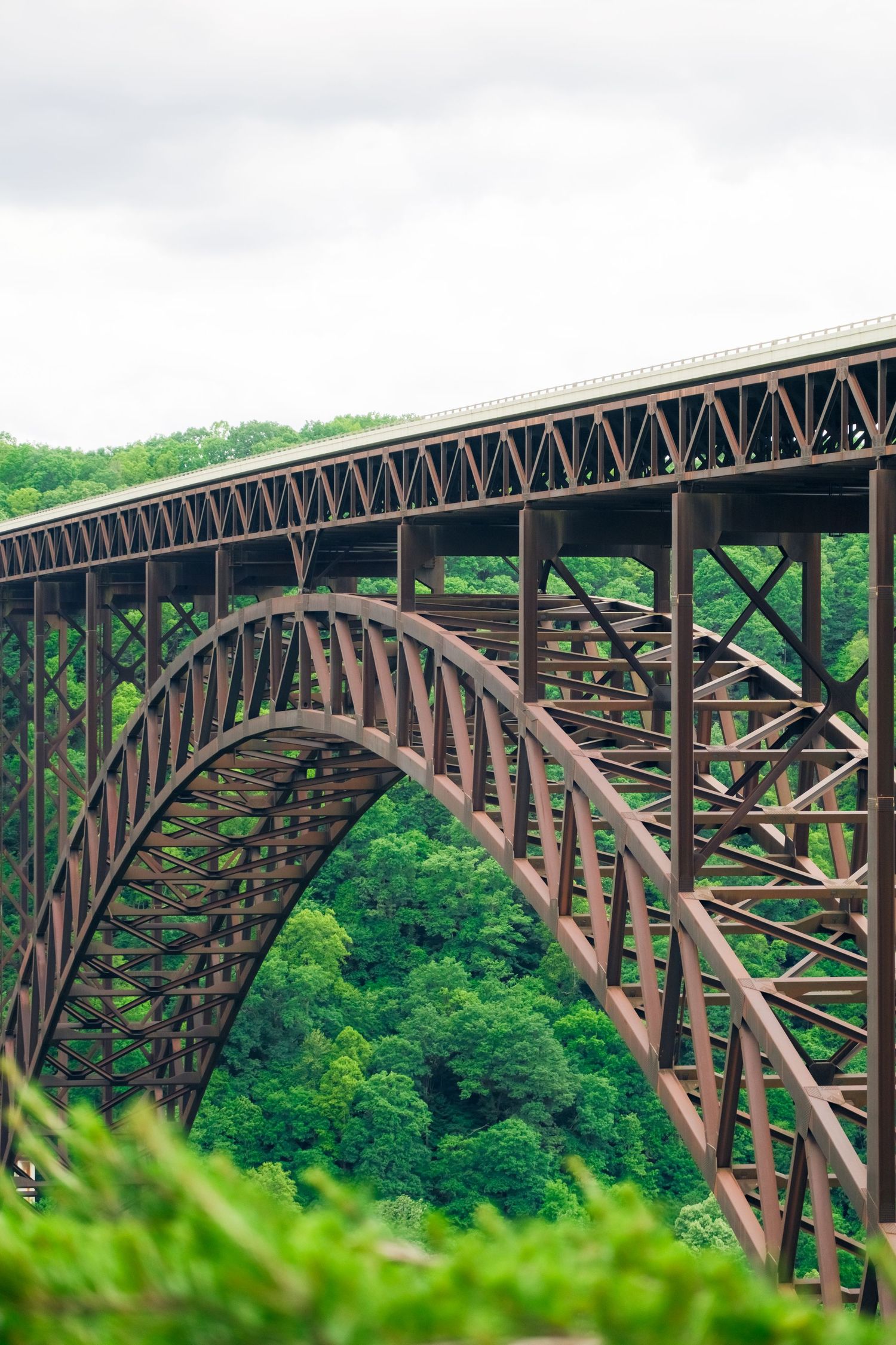 A large rustic steel arch bridge spans across a lush green forested valley with structural trusses visible in its design.