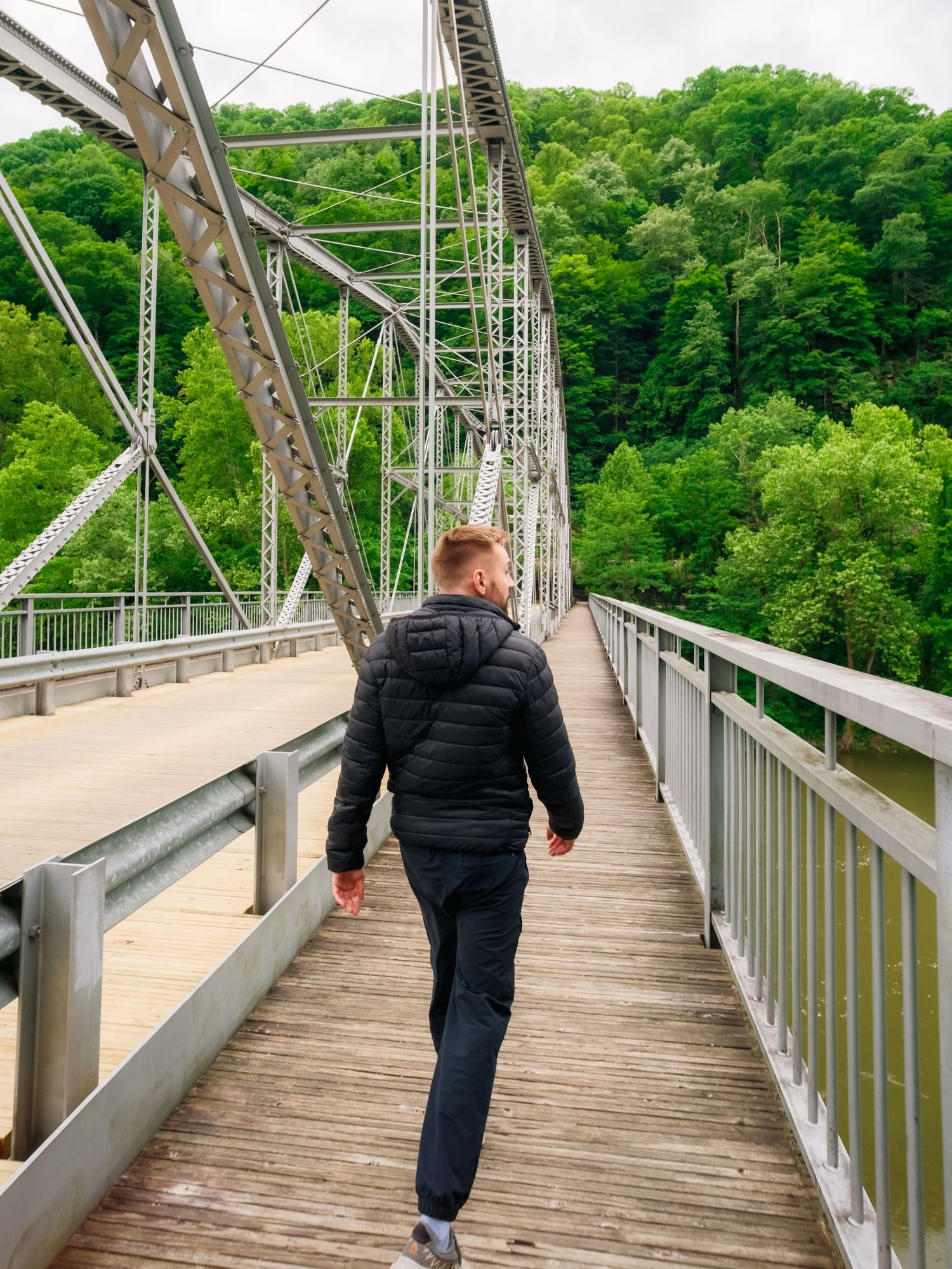 Person walking across a wooden bridge with metal truss architecture surrounded by lush green forest landscape.