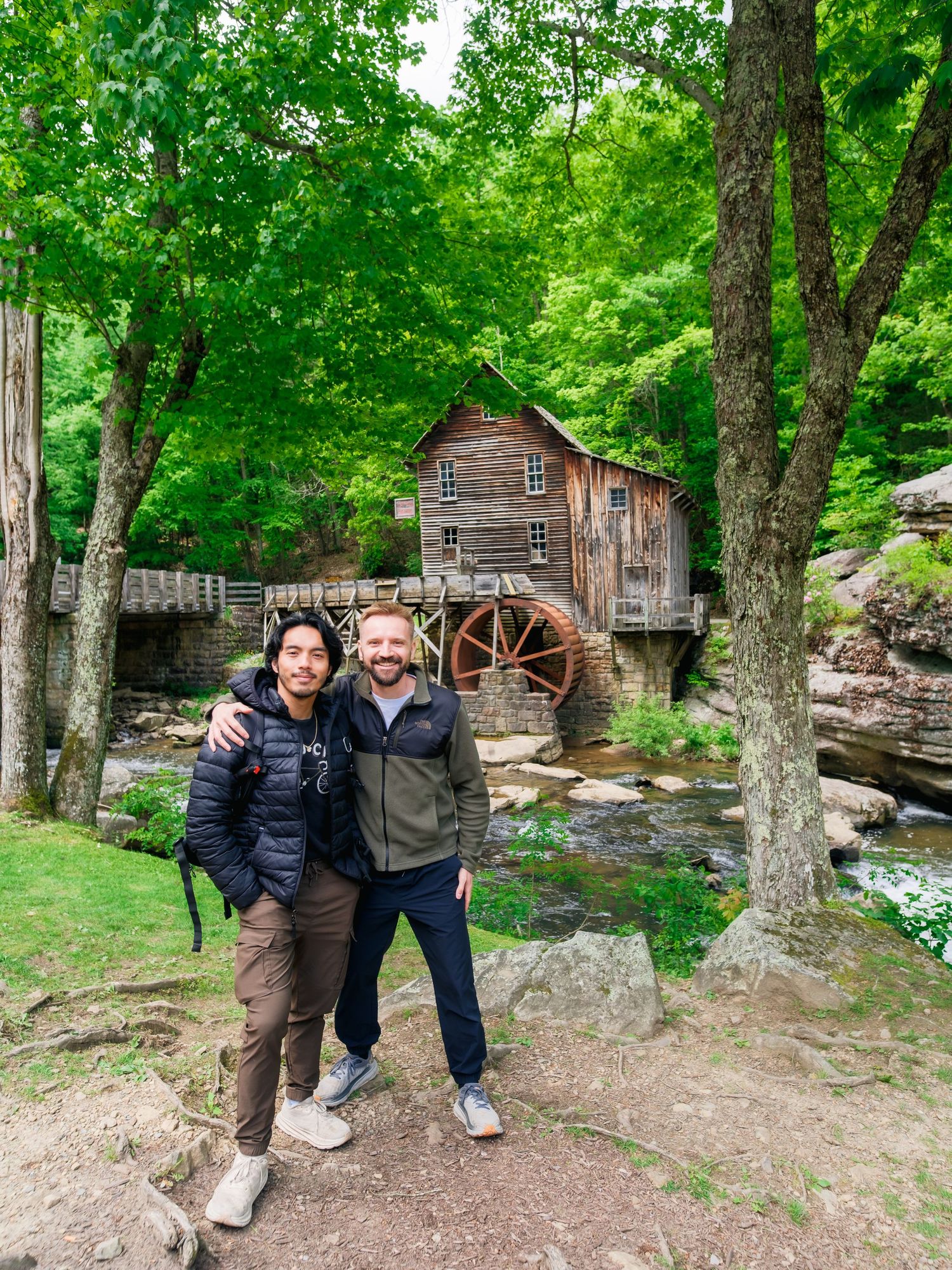 Two visitors stand in front of the historic Glade Creek Grist Mill surrounded by lush green forest at Babcock State Park, West Virginia.