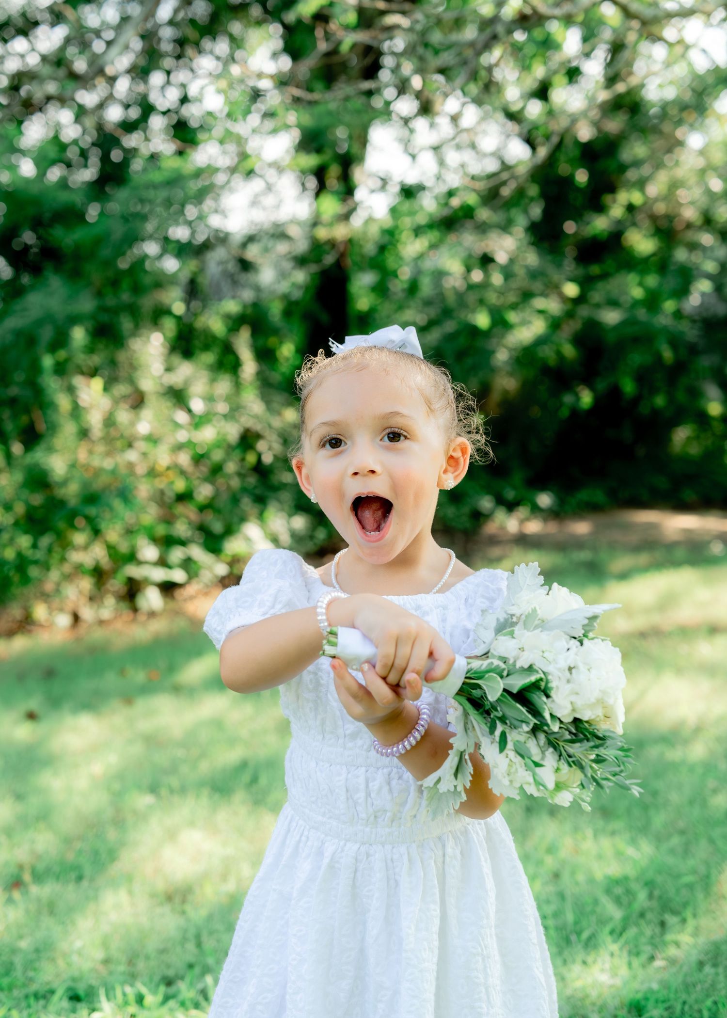 A joyful child in a white dress holds a floral bouquet while making a silly expression in a sunny garden setting.