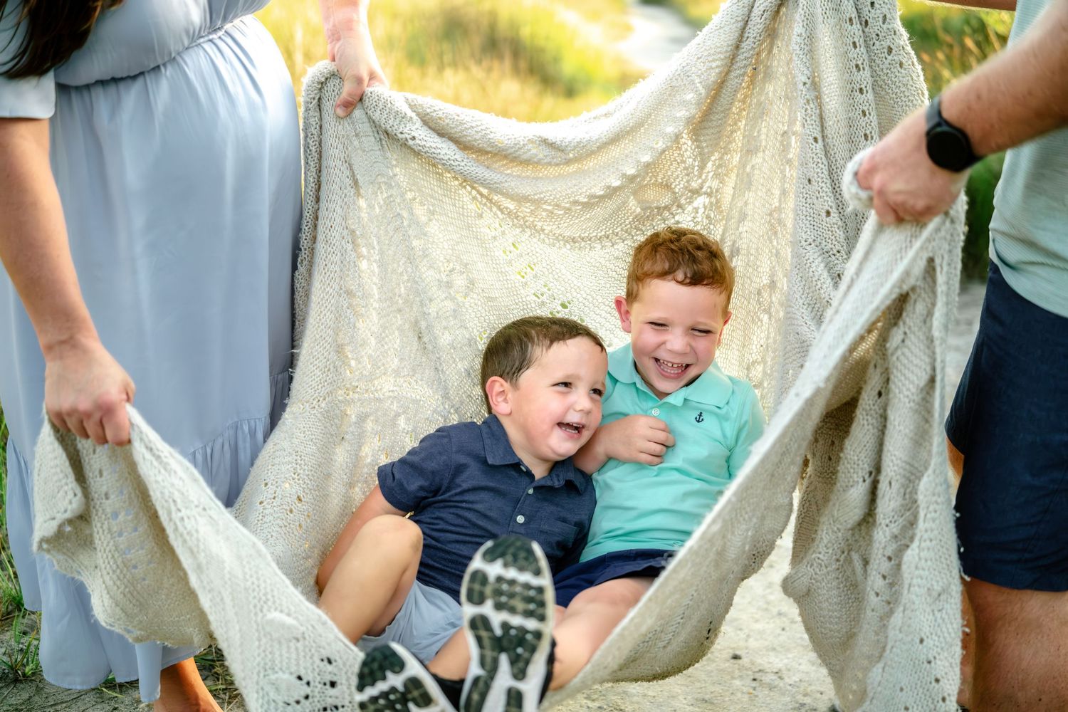 Two children laugh and play in a white blanket being held up by adults outdoors on a sunny day.
