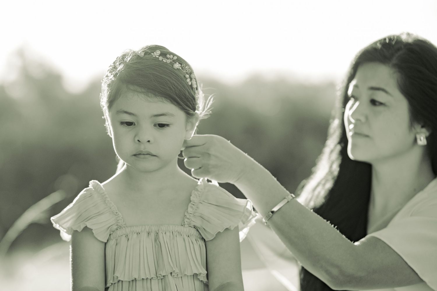 A black and white photo of a woman gently brushing a young child's hair outdoors in soft natural lighting.