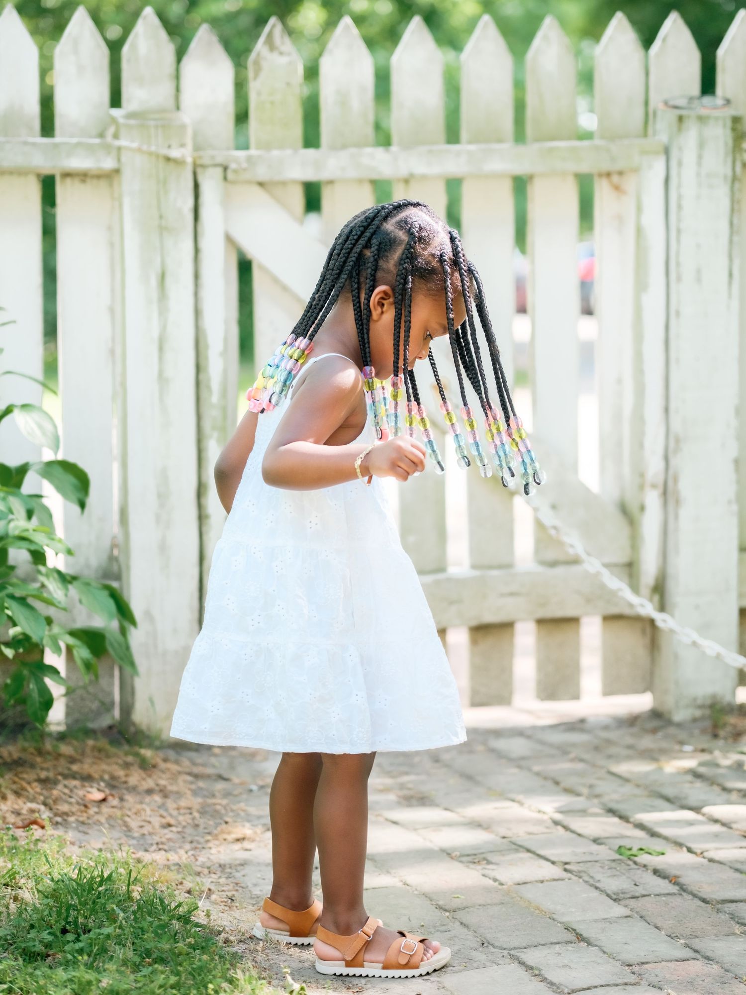 Small child in white summer dress standing on stone path by white picket fence in garden.