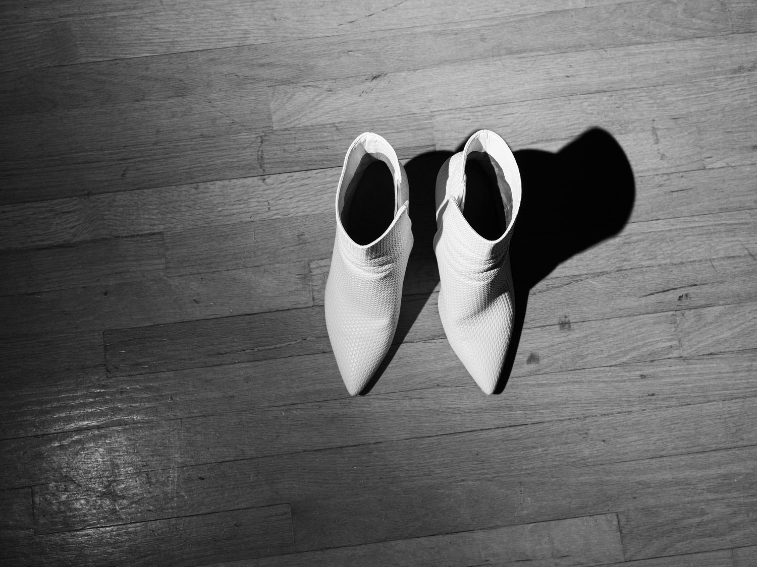 A pair of white leather ankle boots sits on a hardwood floor casting dramatic shadows in black and white.