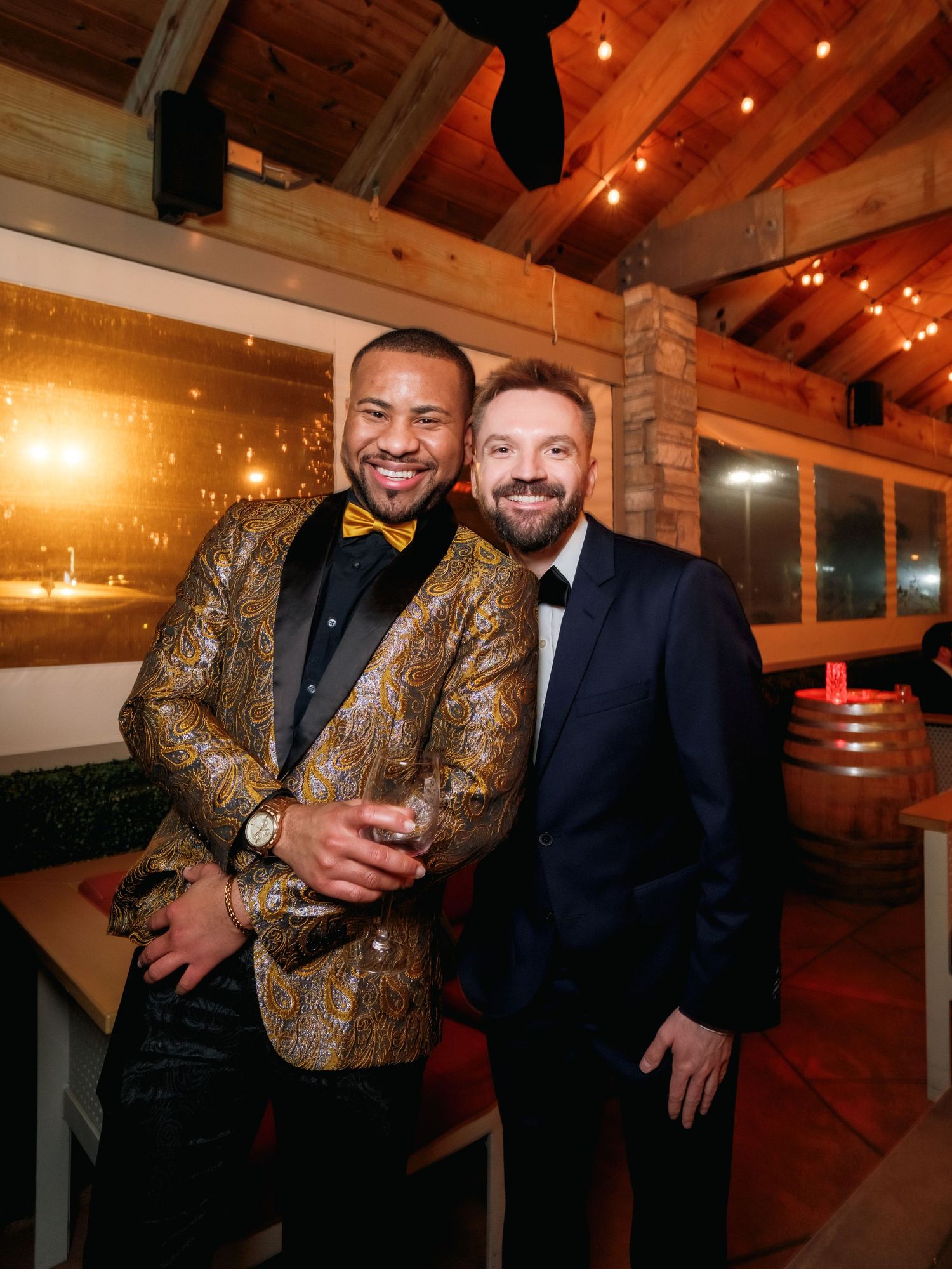 Two guests at a formal event pose together inside a rustic venue with exposed wooden beams and string lights.