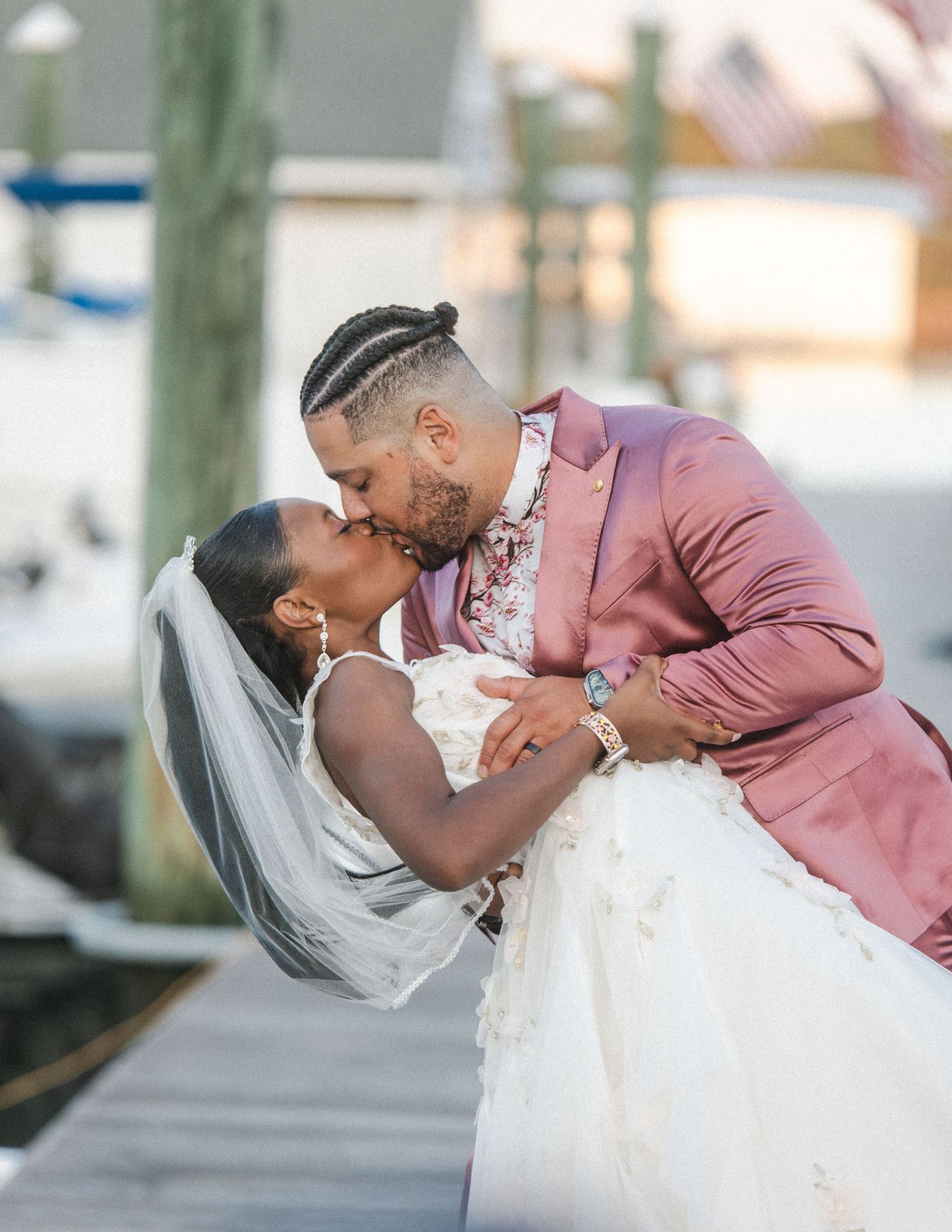 A romantic dip and kiss on a dock at sunset with the person in a mauve suit holding their partner in a white gown.