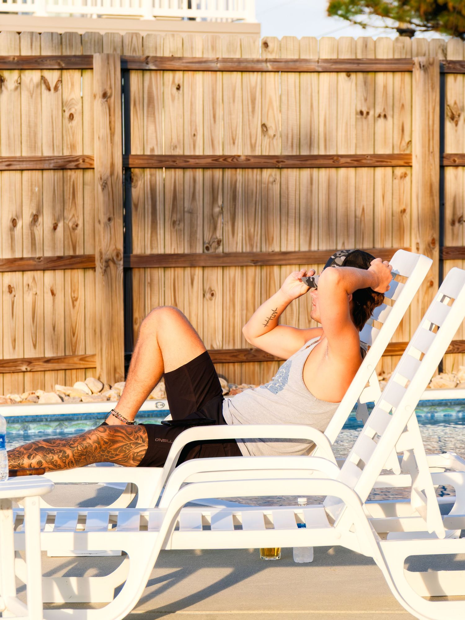 People relaxing on white lounge chairs by a pool during a sunny day at a leisure facility.