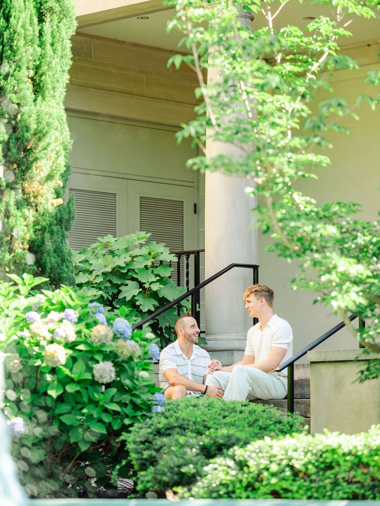 Two people spending a relaxing afternoon on outdoor stairs surrounded by lush green plants and cypress trees.