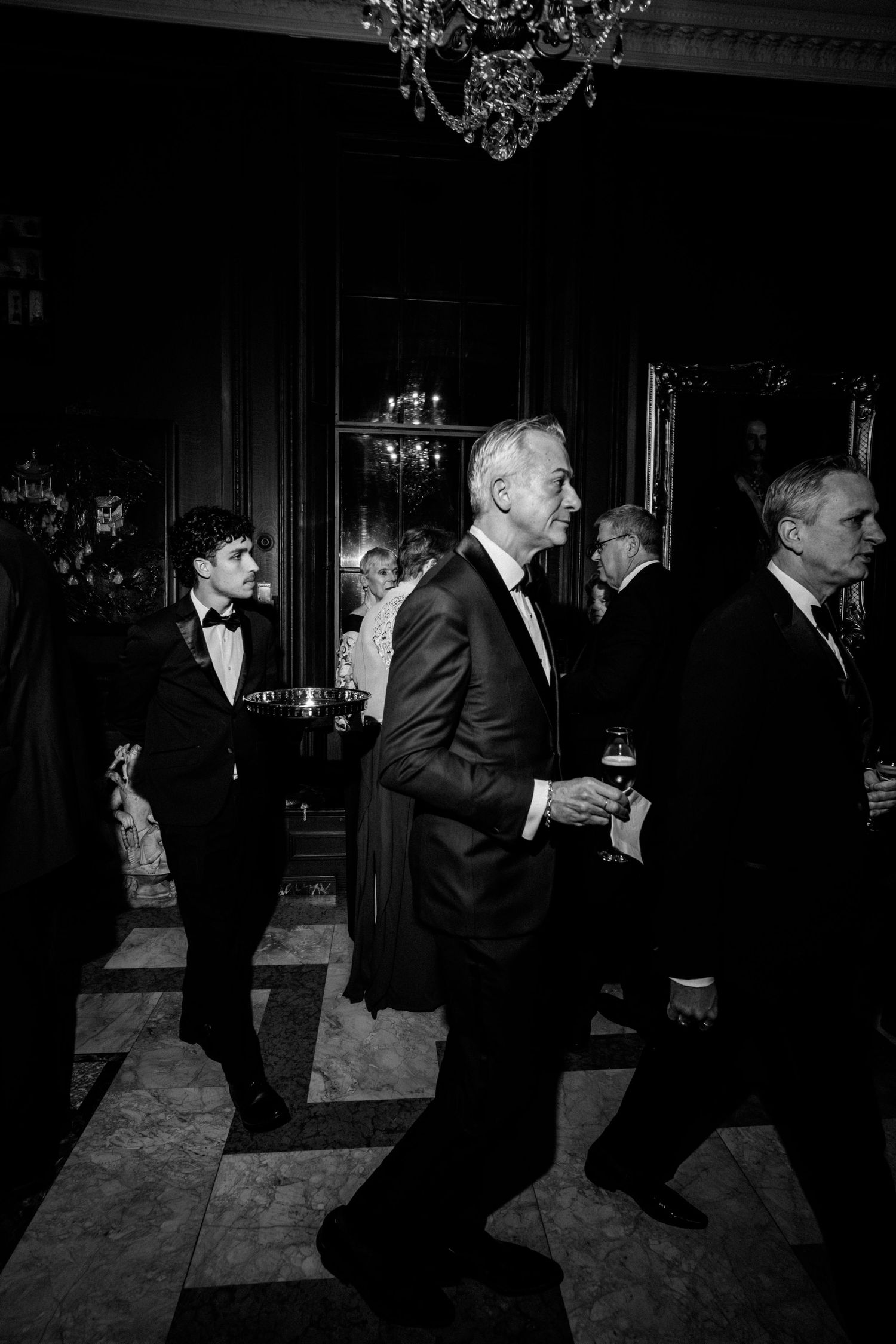 People in formal attire dance together in a dimly lit ballroom with elegant chandeliers overhead.