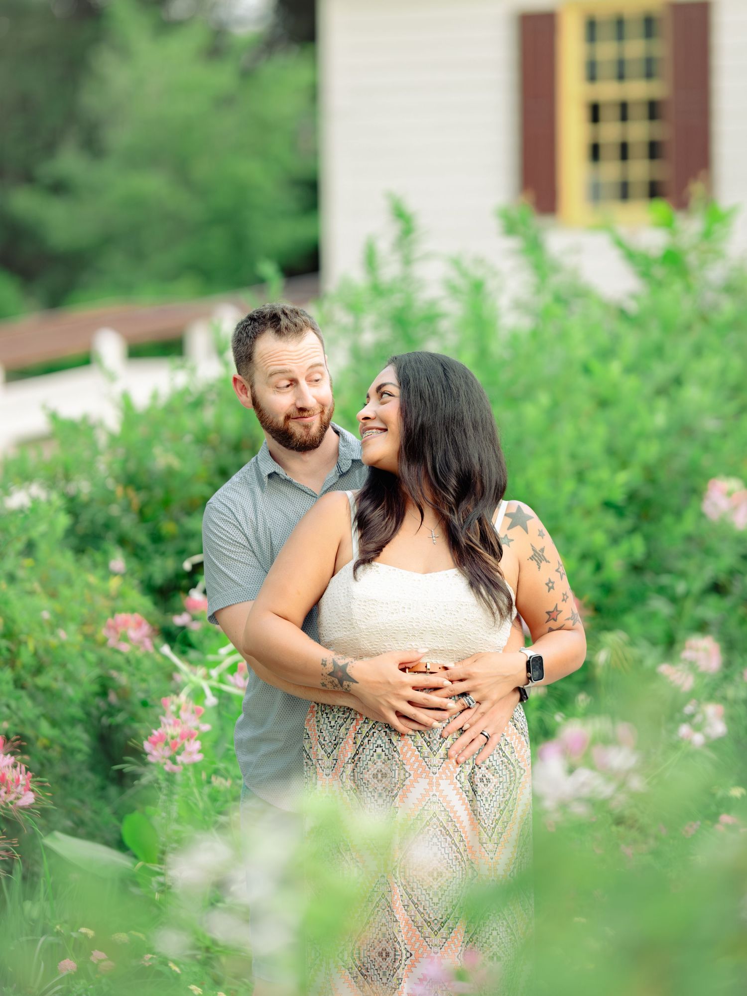 A couple embraces in a lush garden setting with pink flowers blooming around them on a sunny day.