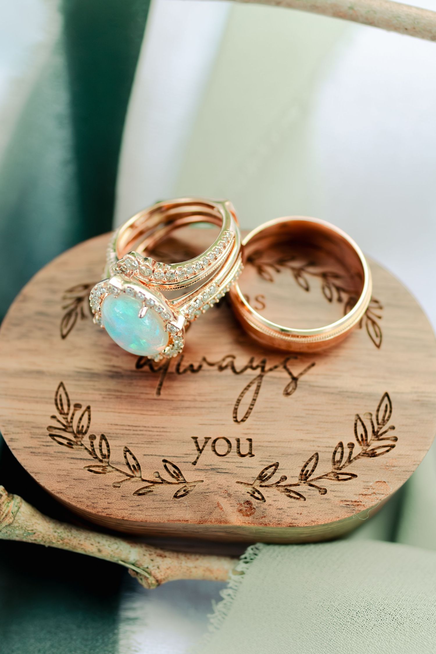 Wedding rings rest on a wooden surface engraved with 'always you' and delicate leaf designs, featuring a rose gold opal ring set.