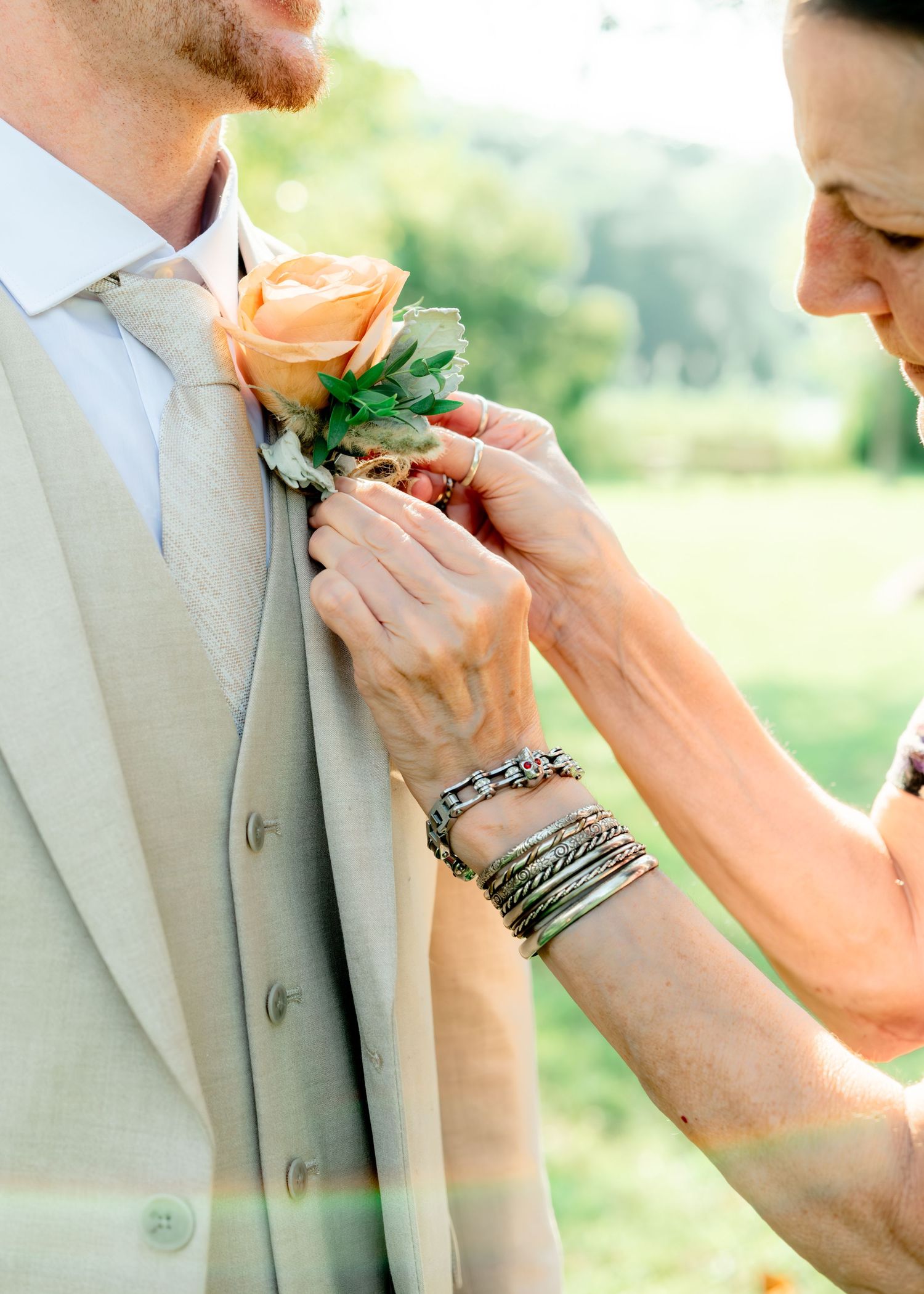Hands carefully pin a peach rose and succulent boutonniere onto a light gray suit jacket at an outdoor wedding.