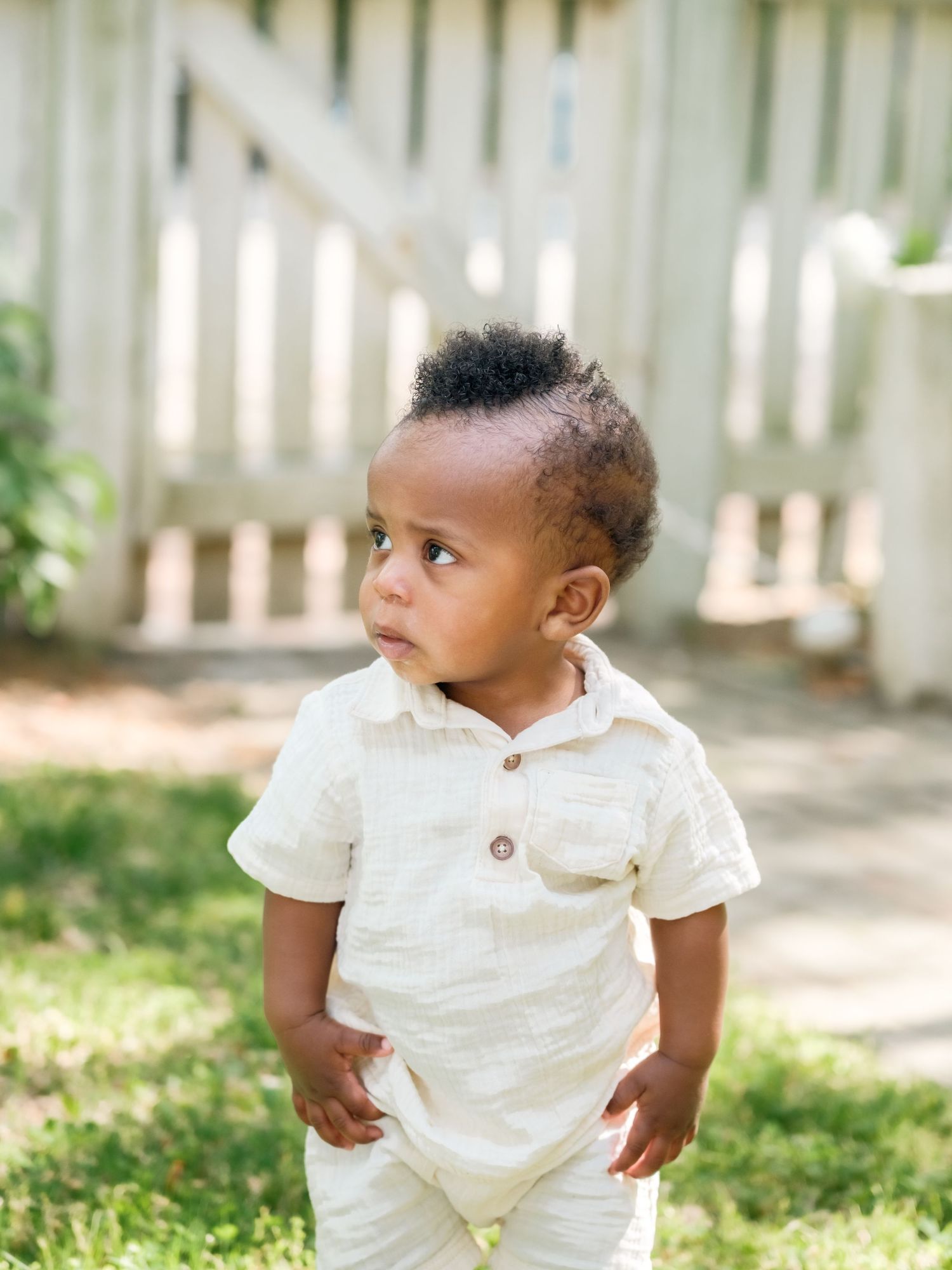 Small child wearing a white collared shirt and light pants stands on grass in front of a white picket fence.