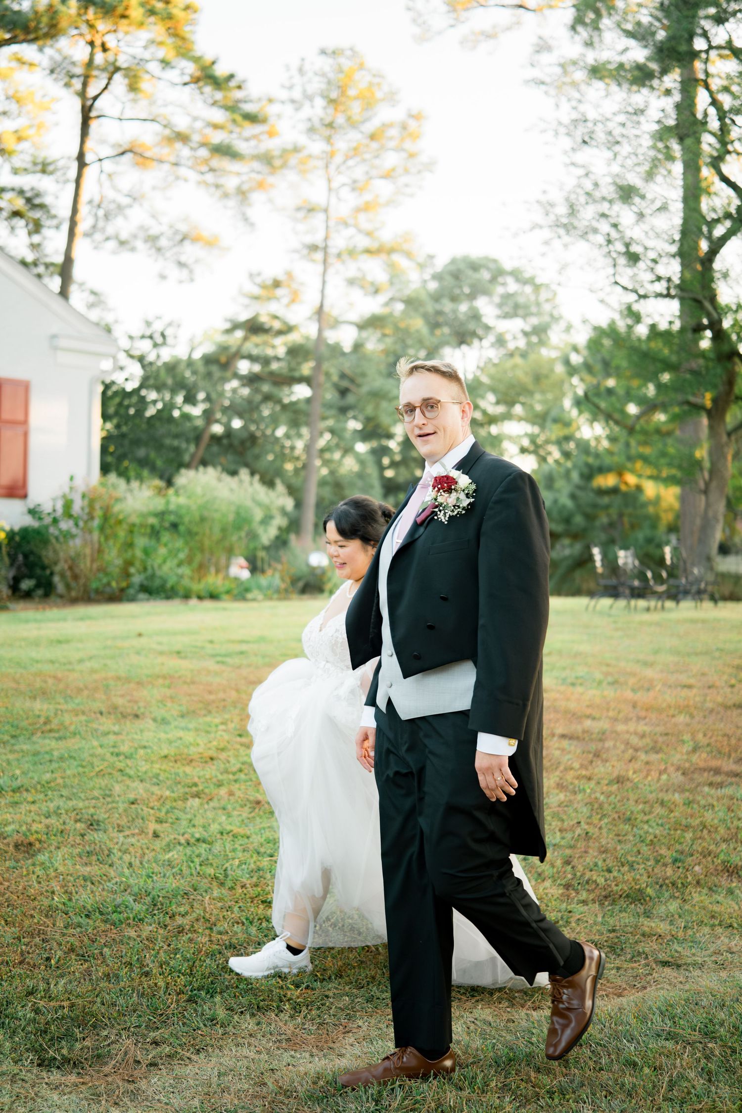 A couple in wedding attire walks across a grassy lawn near a white building with tall trees in the background.