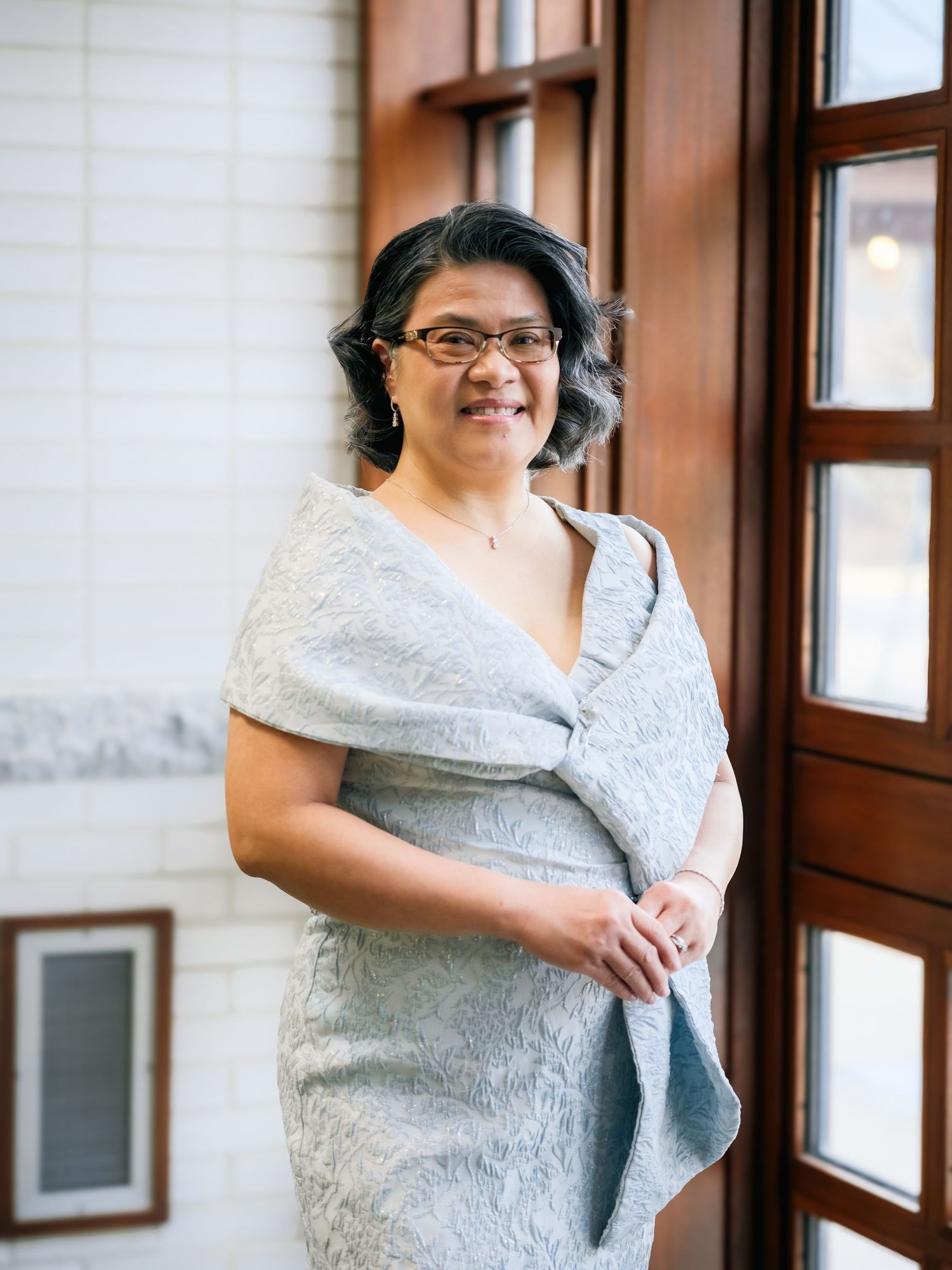 Person in white lace dress and glasses stands by window in elegant indoor setting.