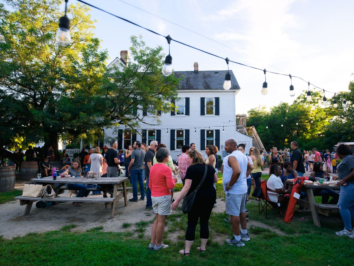 People gather outdoors at a backyard party with string lights and white house in the background during sunset.