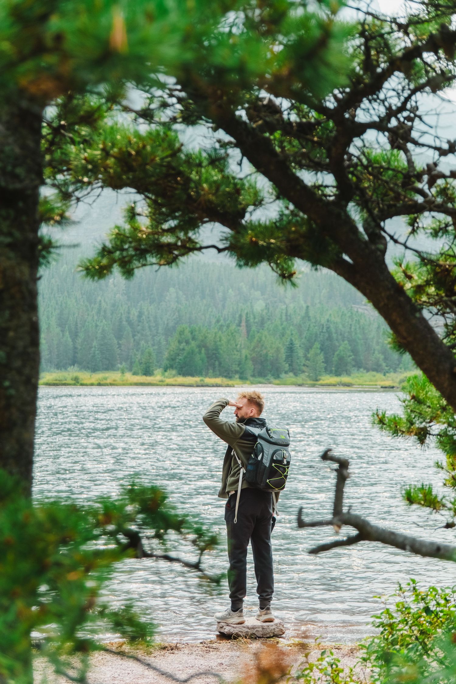 Hiker with backpack overlooking a serene mountain lake surrounded by evergreen forest on a misty day.