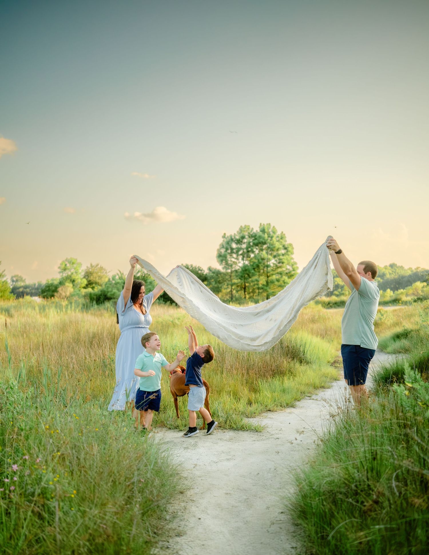 A flowing white veil billows in the breeze along a winding dirt path through tall summer grasses at sunset.