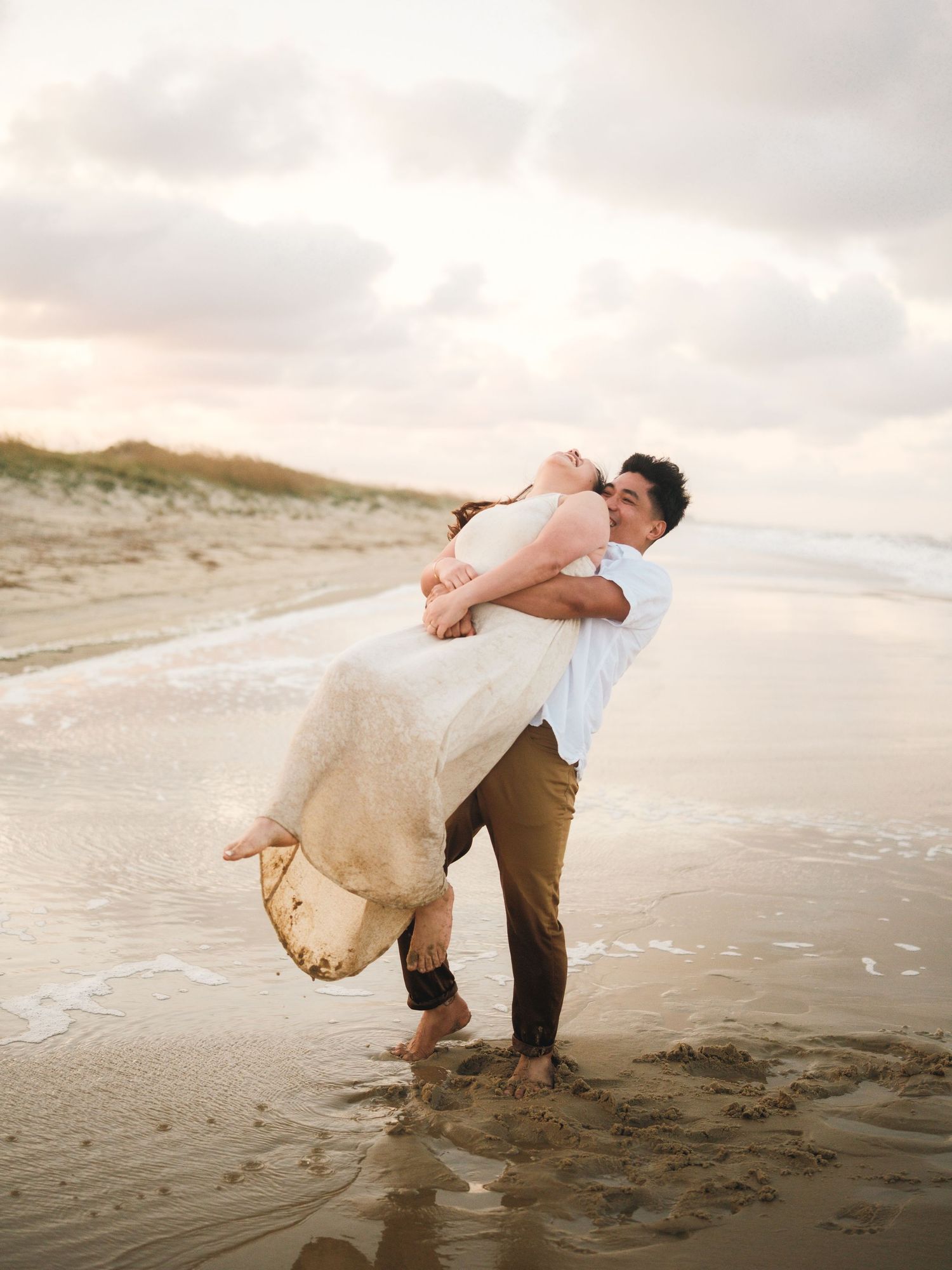 A romantic couple dances and twirls on a sandy beach during golden hour with soft clouds in the sky.