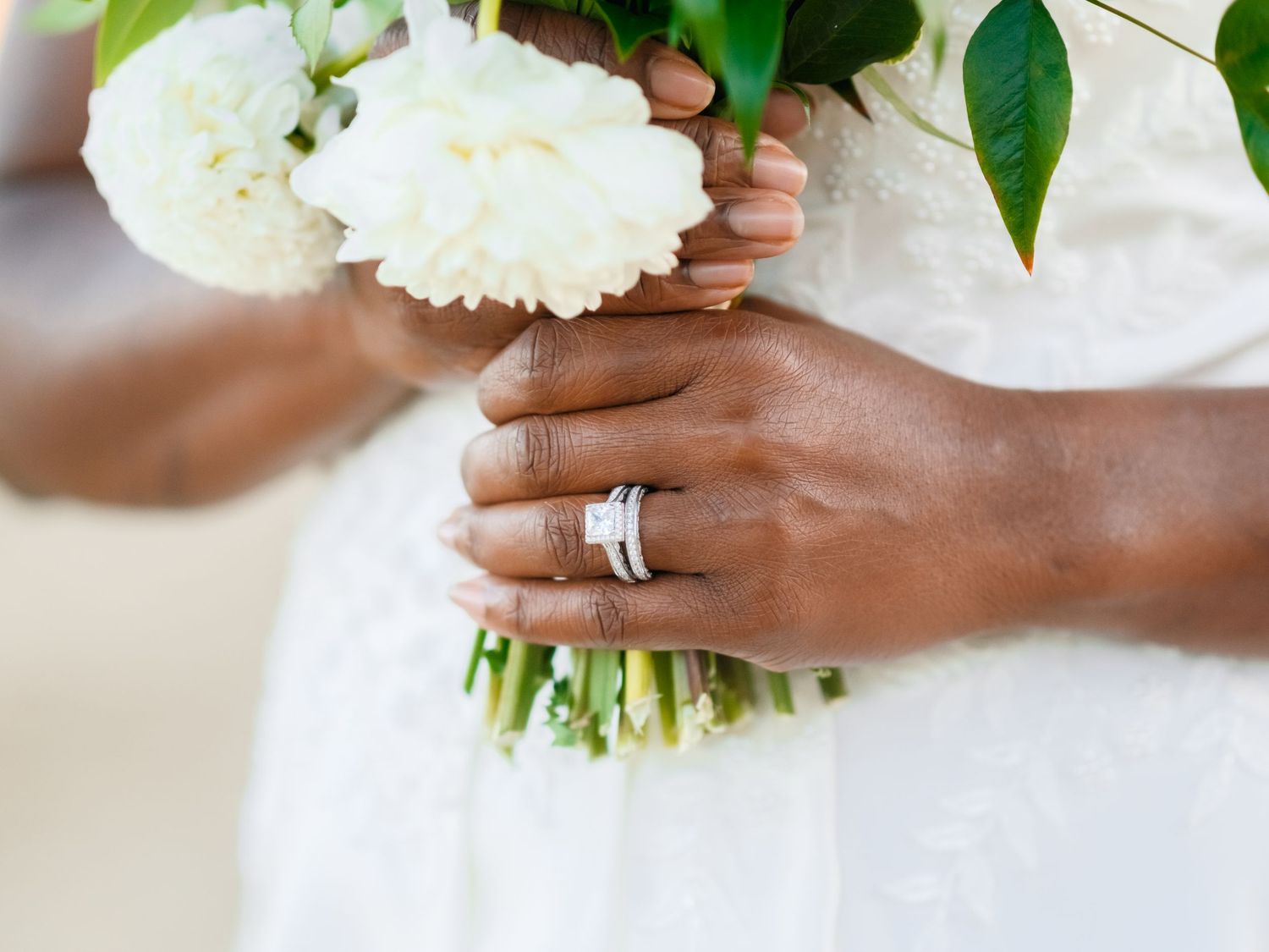 A close-up of hands holding white peonies and wearing a silver wedding band against a light background.