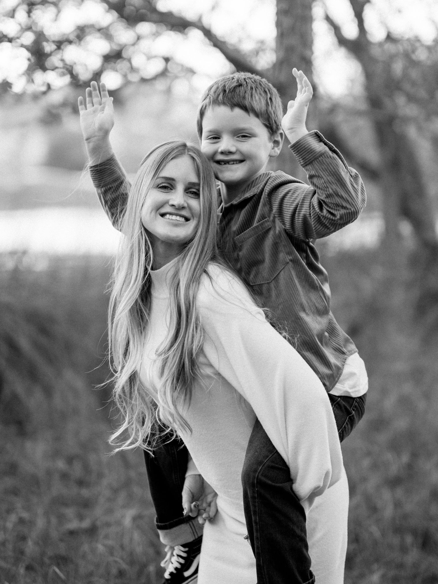 Black and white photo of playful outdoor piggyback ride moment with joyful expressions and raised hands.