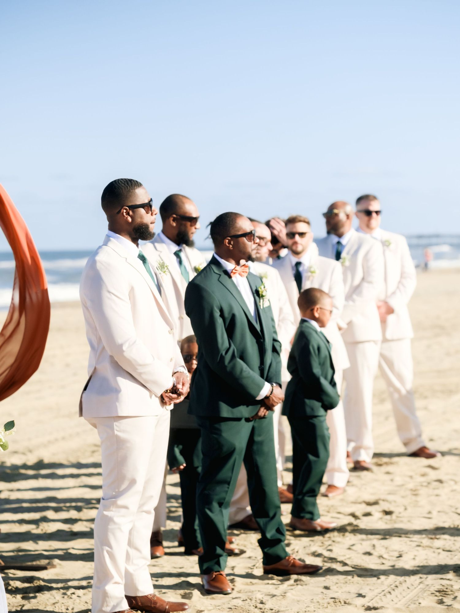 Wedding party members in white and dark green suits stand together on a sunny beach.
