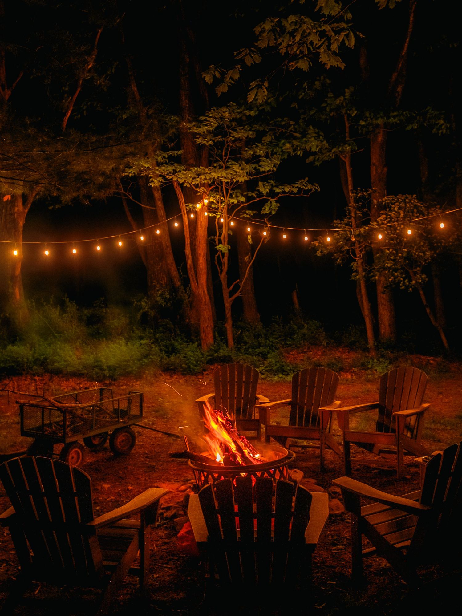 Cozy outdoor scene with Adirondack chairs around a glowing campfire under string lights and trees at night.