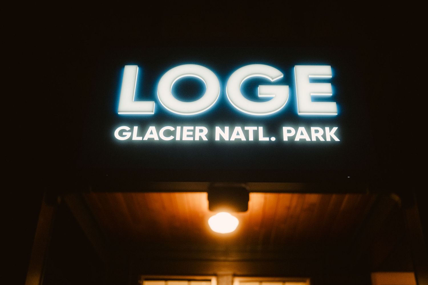 Illuminated lodge sign and entrance at Glacier National Park glows warmly against dark night sky.