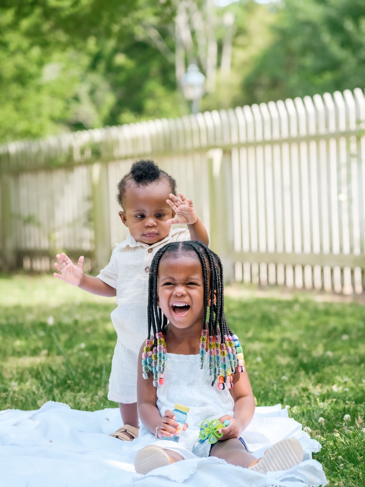 Two children sitting on a white blanket in sunny backyard with picket fence, one laughing with colorful braided hair.
