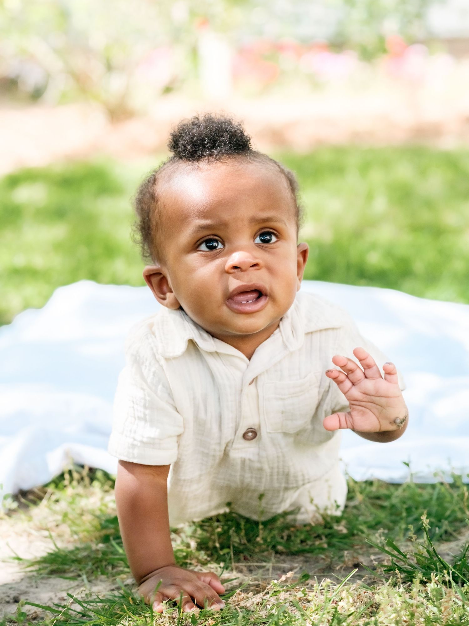 A baby in a cream-colored sweater crawls on grass while smiling during an outdoor photo session.