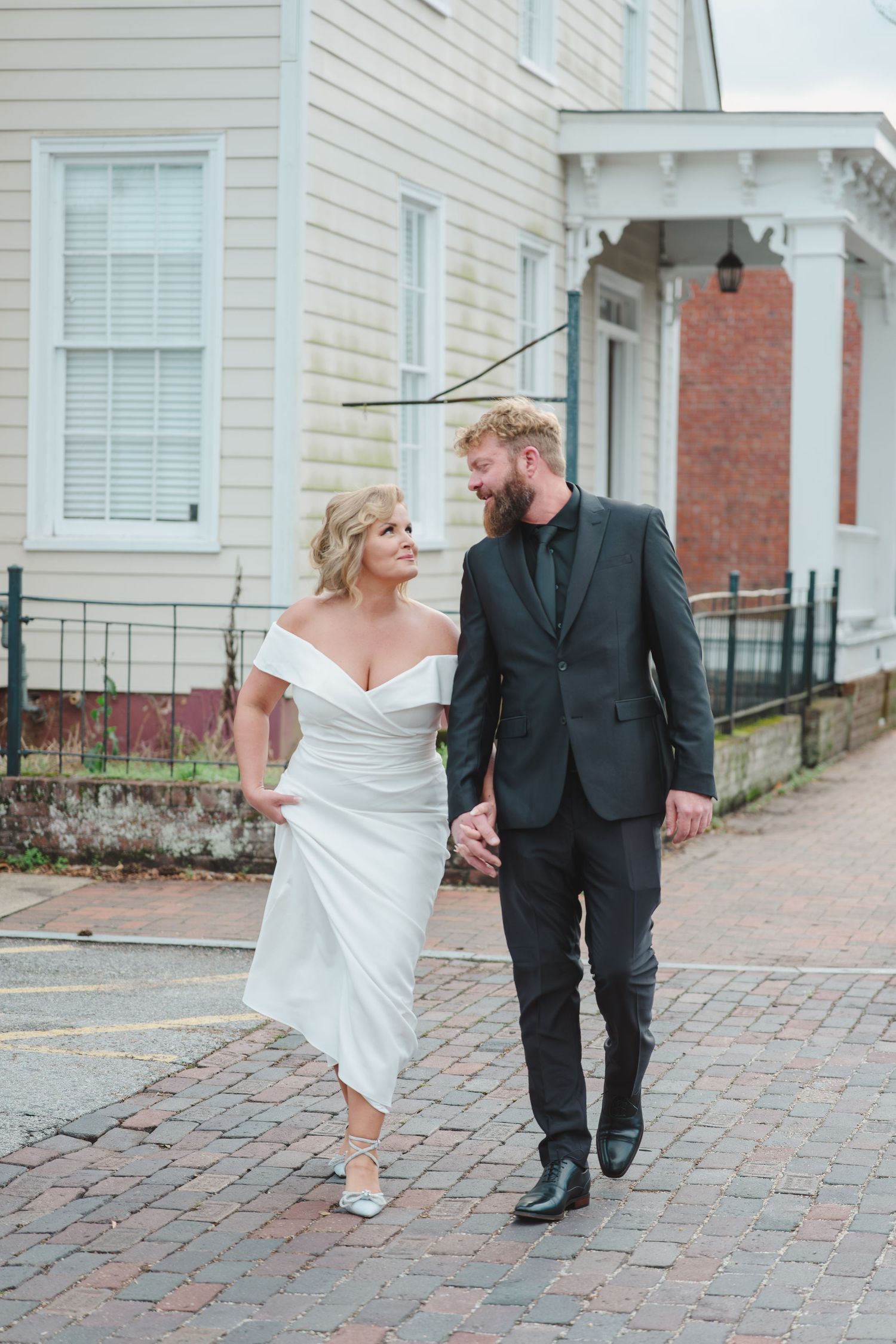 A couple walks together on a brick sidewalk, the bride in an off-shoulder white gown and groom in a dark suit.